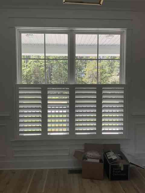 White window with shutter blinds in a room, with boxes on the floor and greenery outside.