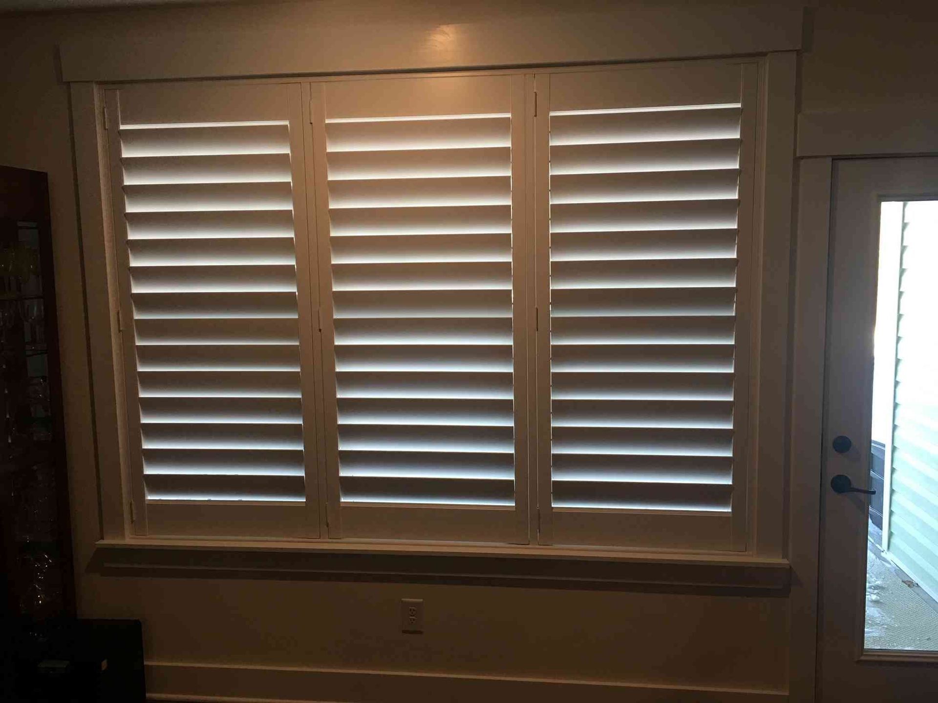 Three white window shutters on a beige wall beside a glass door with bright light outside