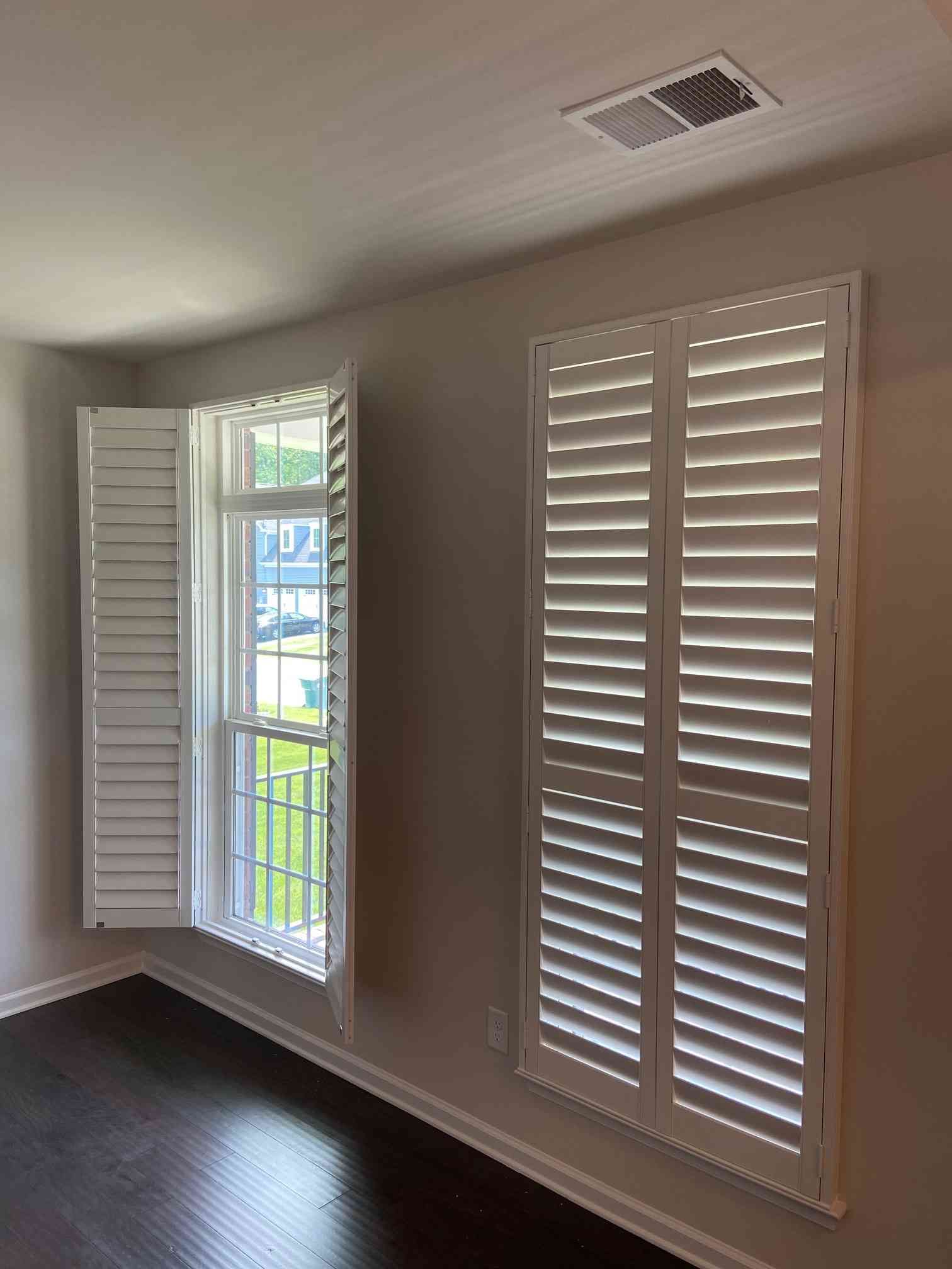 Empty room with dark wood floors, beige walls, and two white shuttered windows, one open to a balcony.