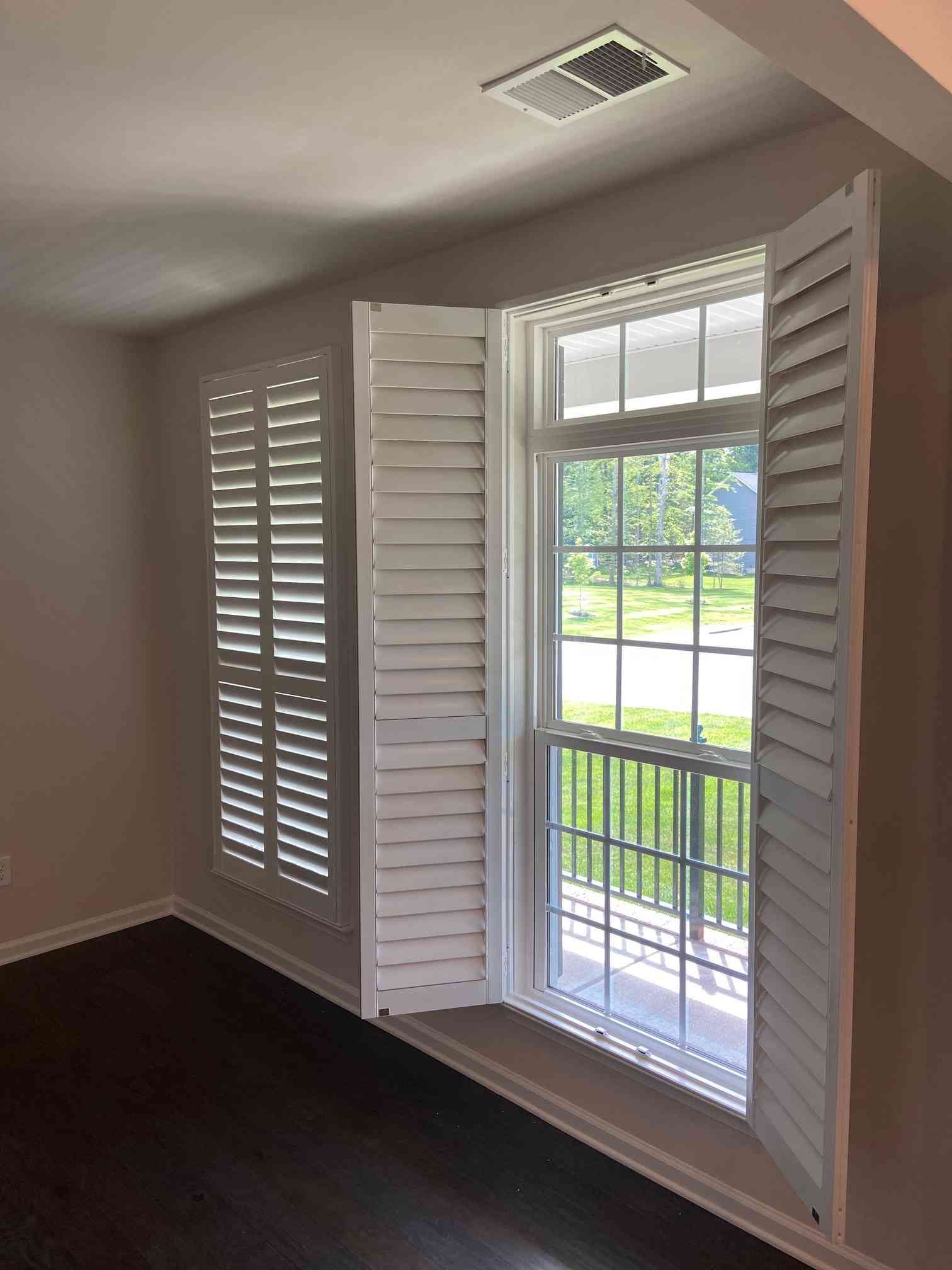 Bright room with white shutters open to a balcony and green yard outside, dark wood floor and ceiling vent.