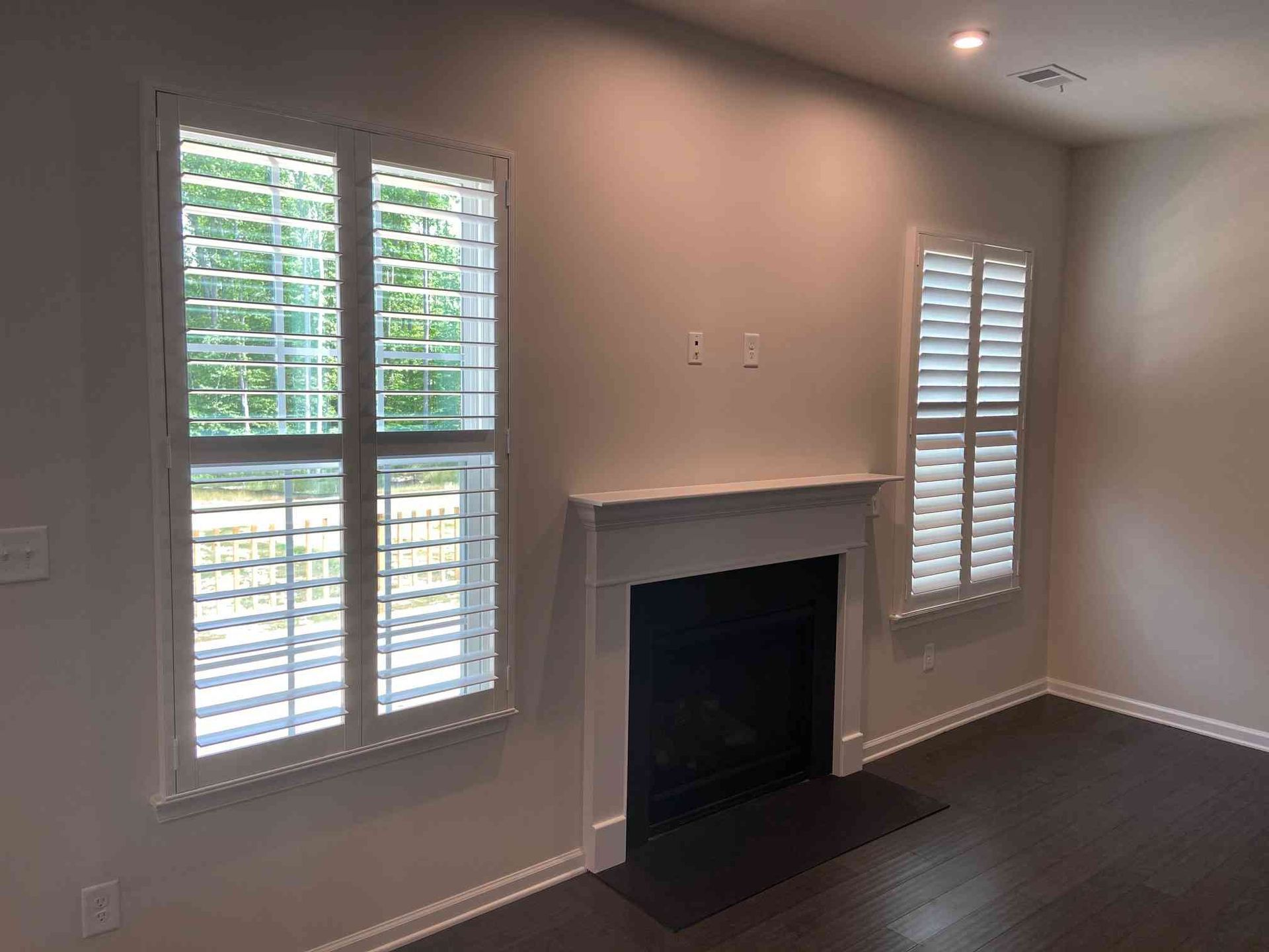 Empty living room with fireplace, two windows, beige walls, and white plantation shutters.