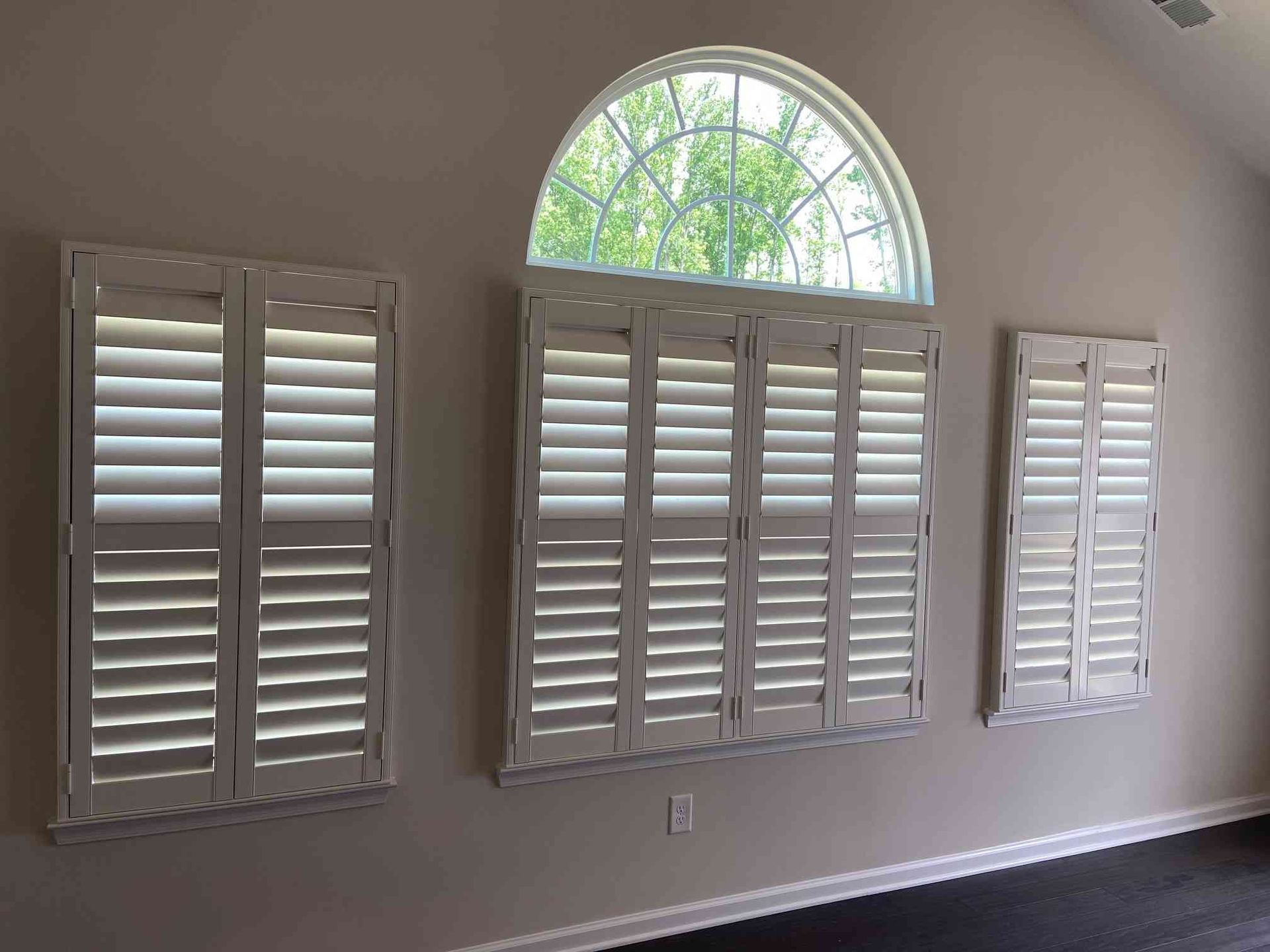Three white plantation shutters on a beige wall beneath an arched window.