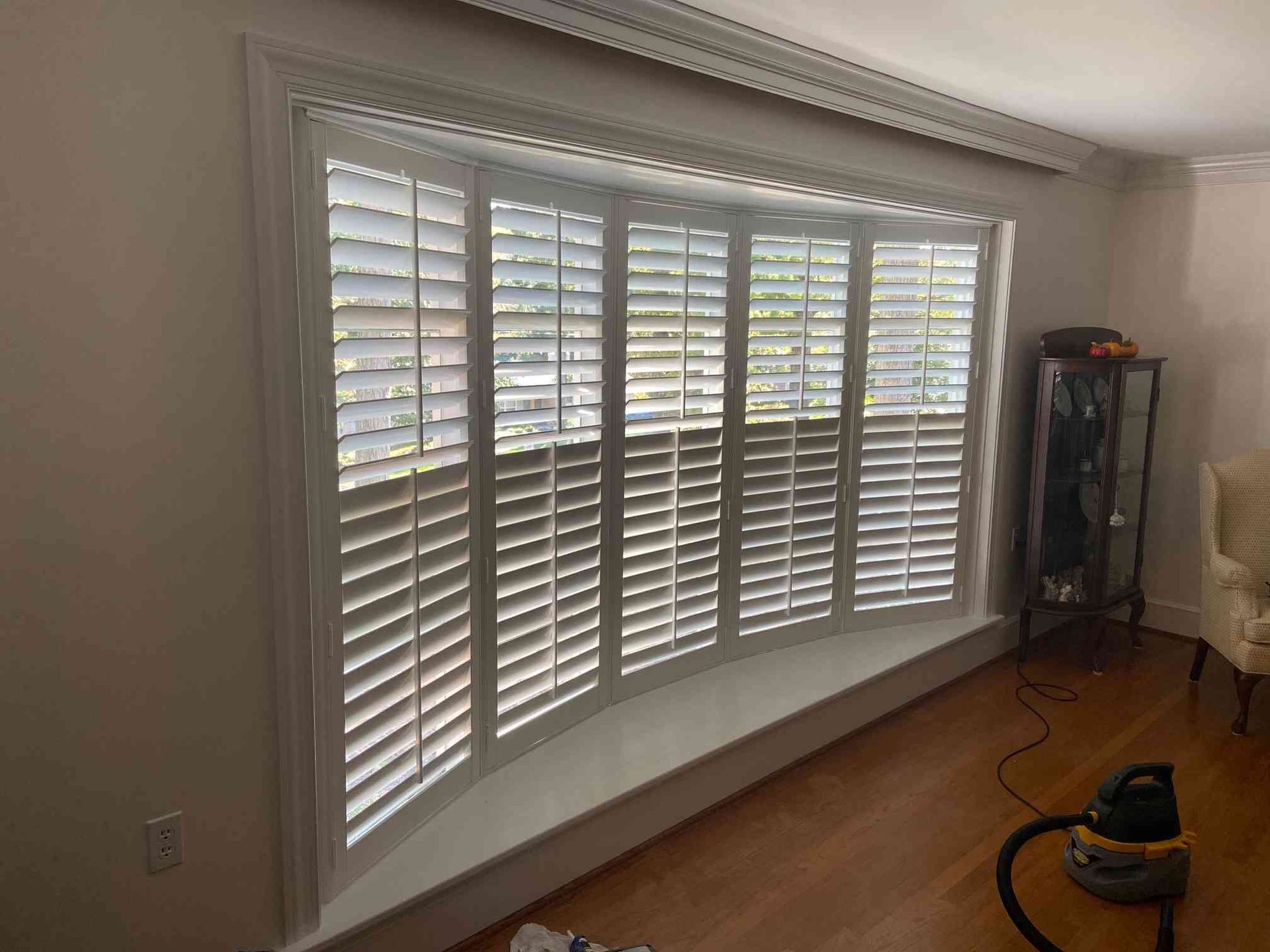 Bright bay window with white plantation shutters in a living room; vacuum on hardwood floor.