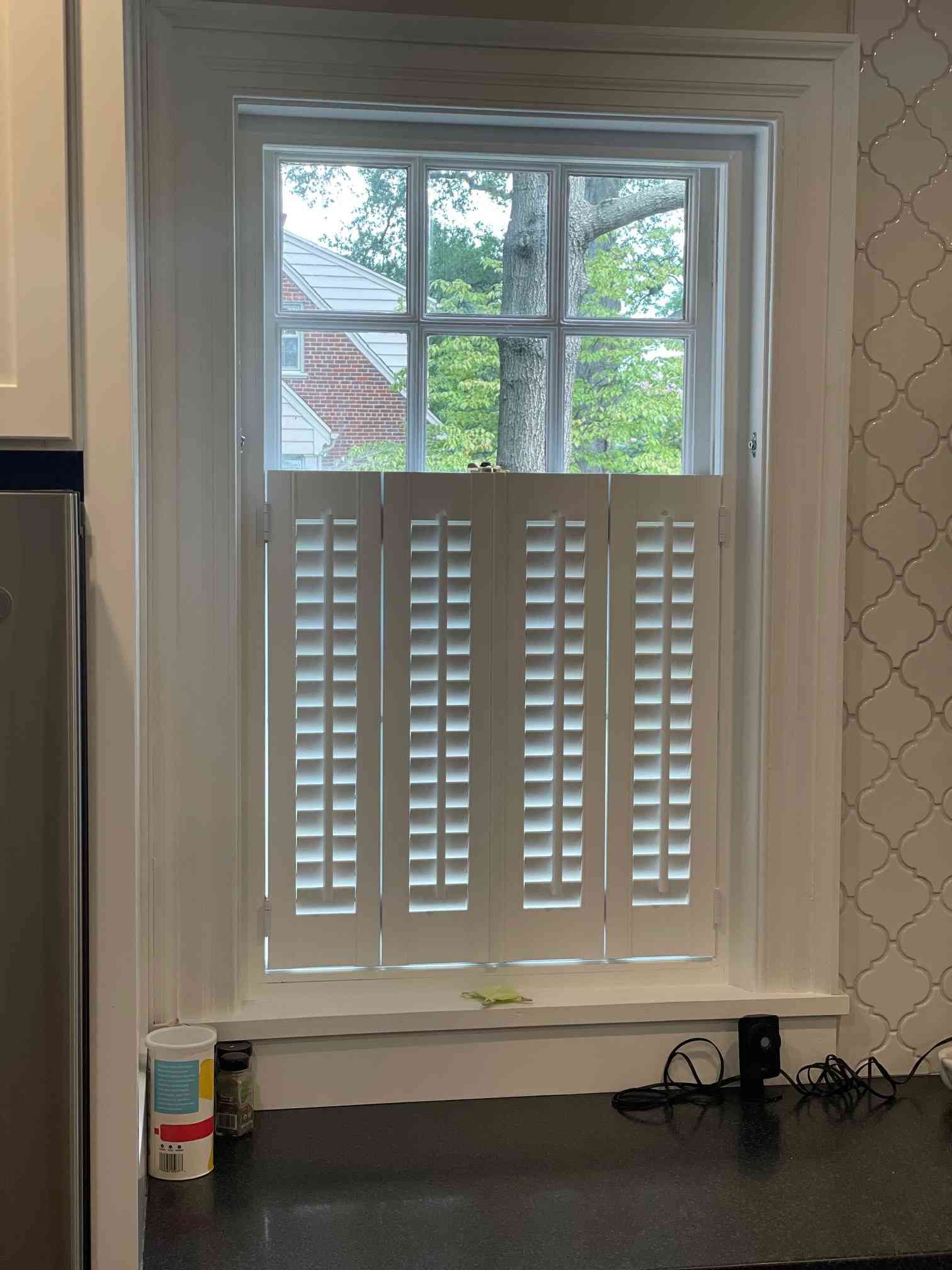 Kitchen window with white shutters and a black countertop below, overlooking trees and neighboring houses
