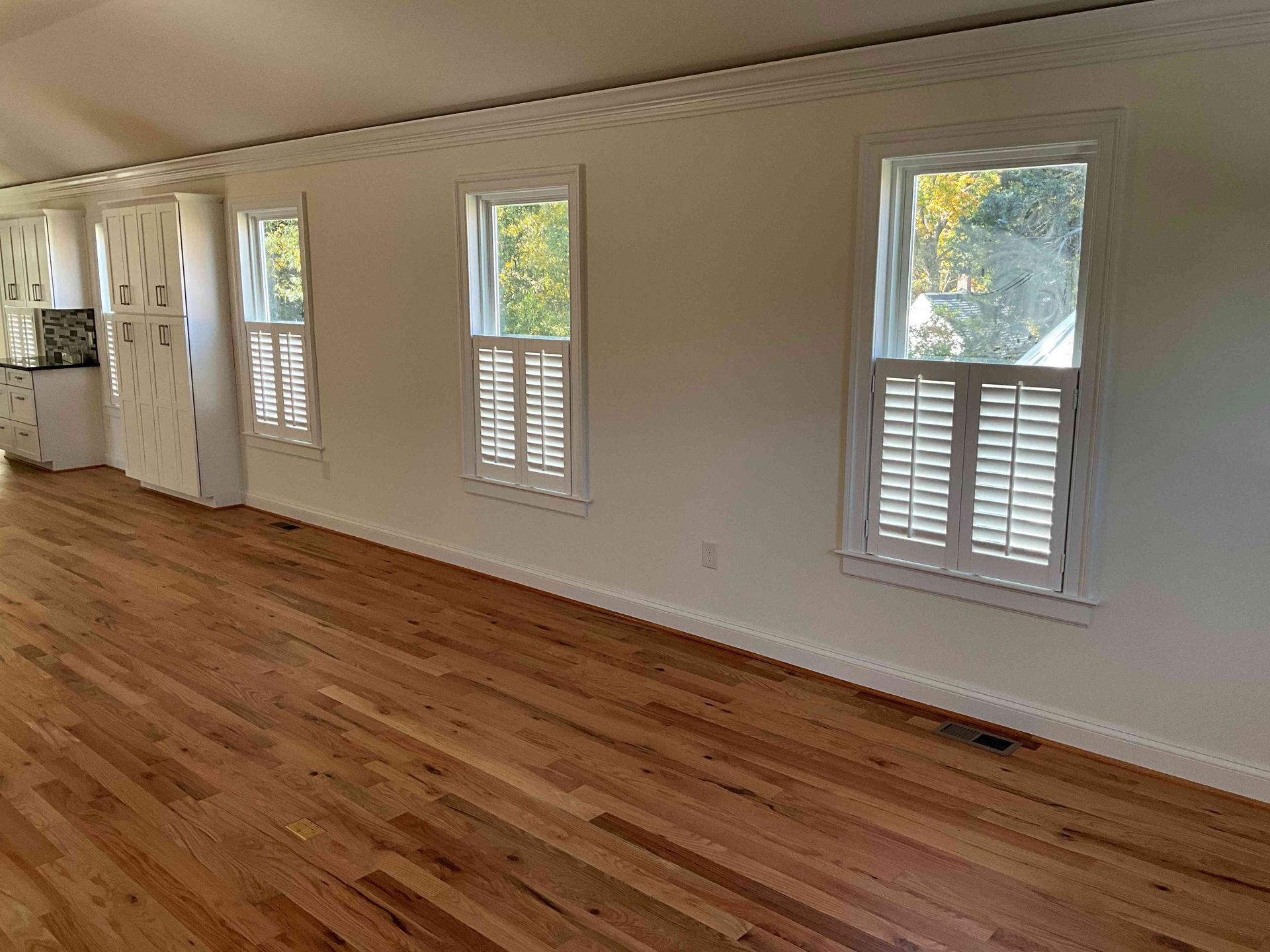 Empty room with hardwood floors, white walls, and several tall windows with shutters.