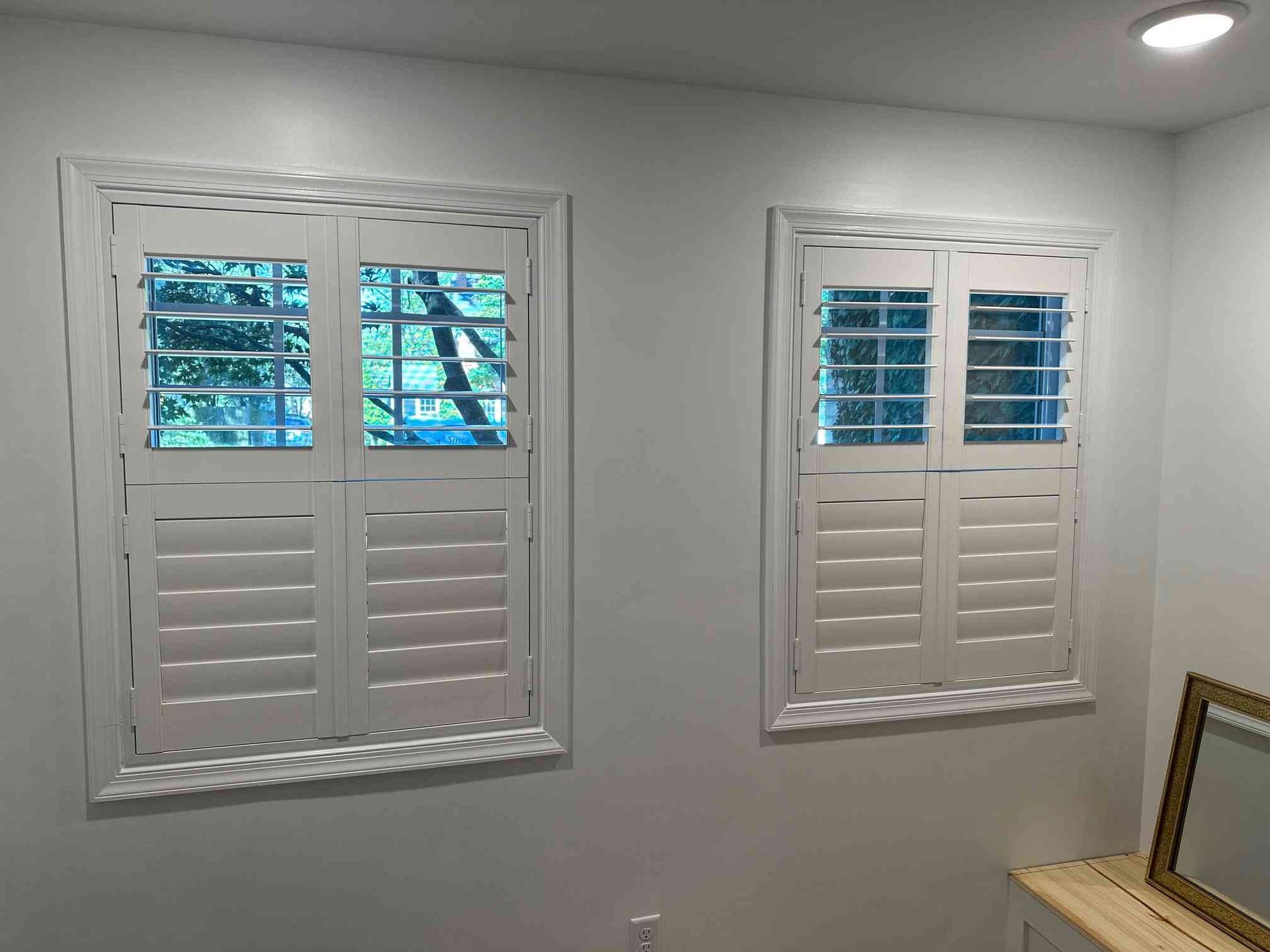 Two white shuttered windows on a light gray wall with a ceiling light above and framed art in the corner