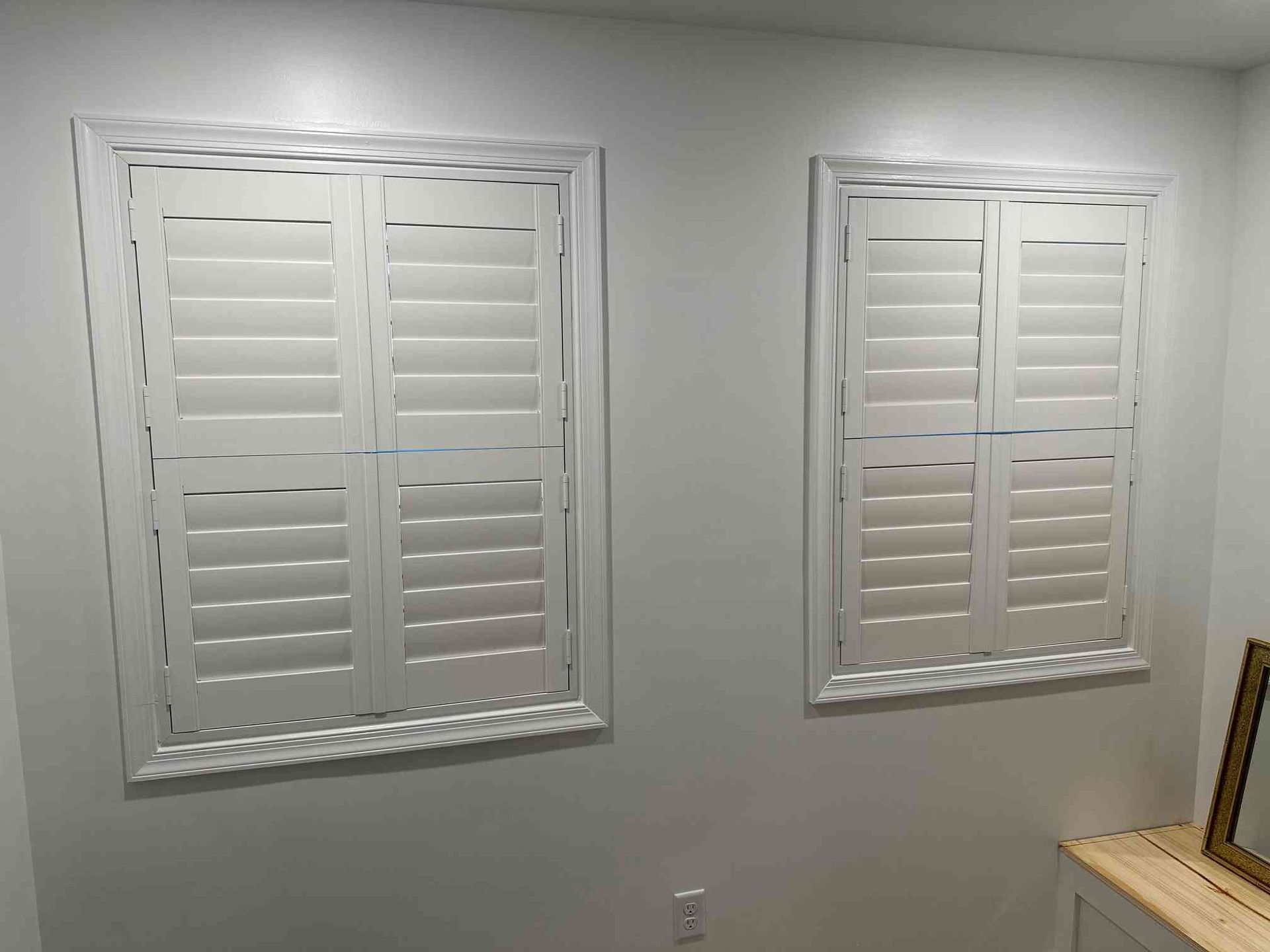 Two white plantation shutters on a light gray wall above a beige chair rail and outlet