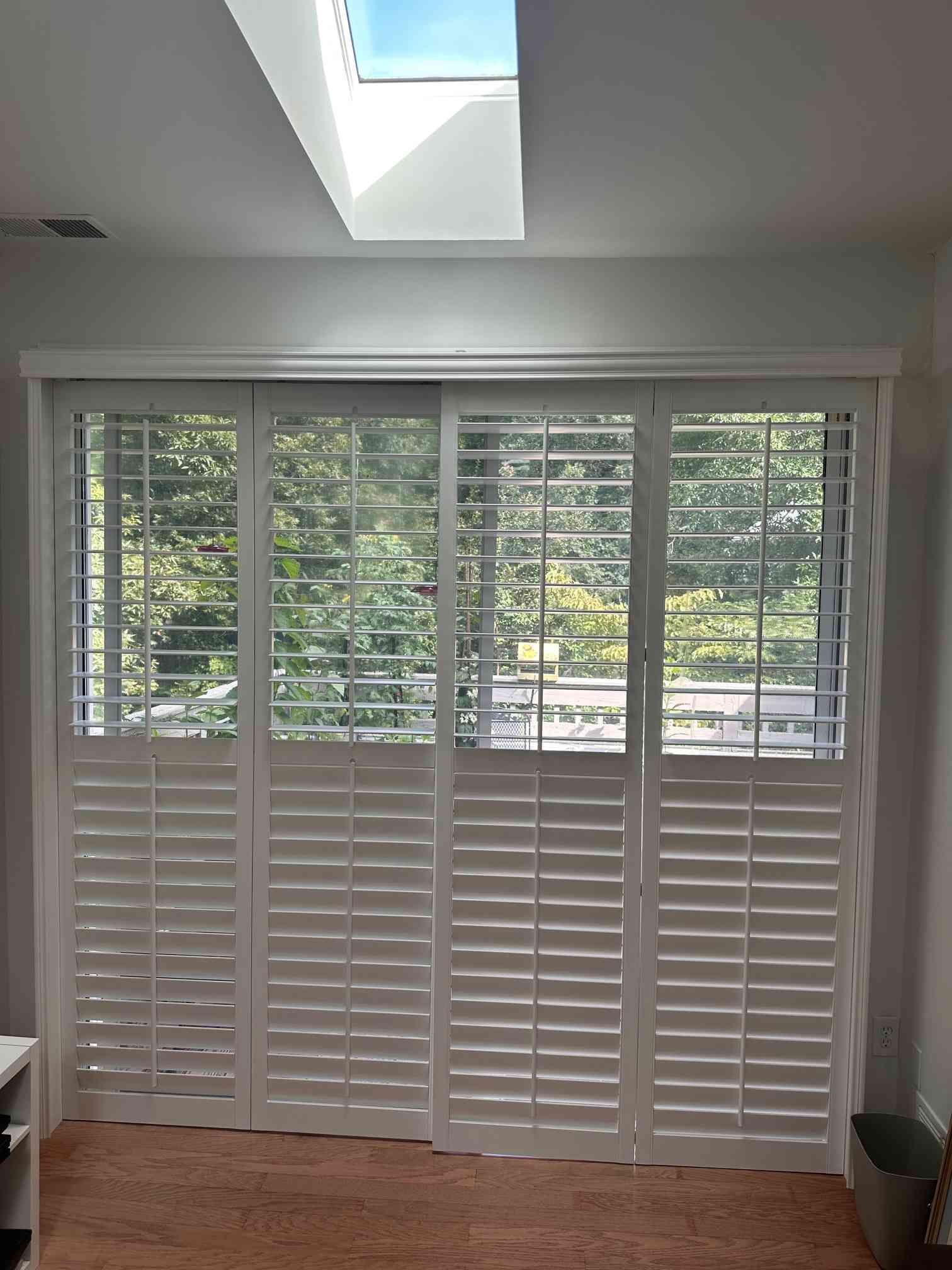 White plantation shutters covering a large window beneath a skylight, with trees visible outside