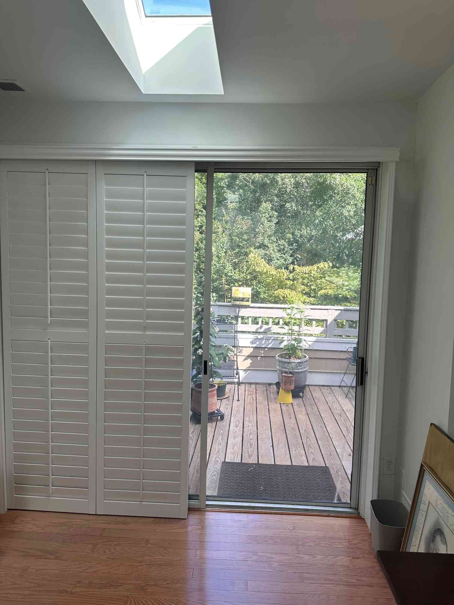Bright room with white shutters opening to a deck and green backyard through sliding glass doors.