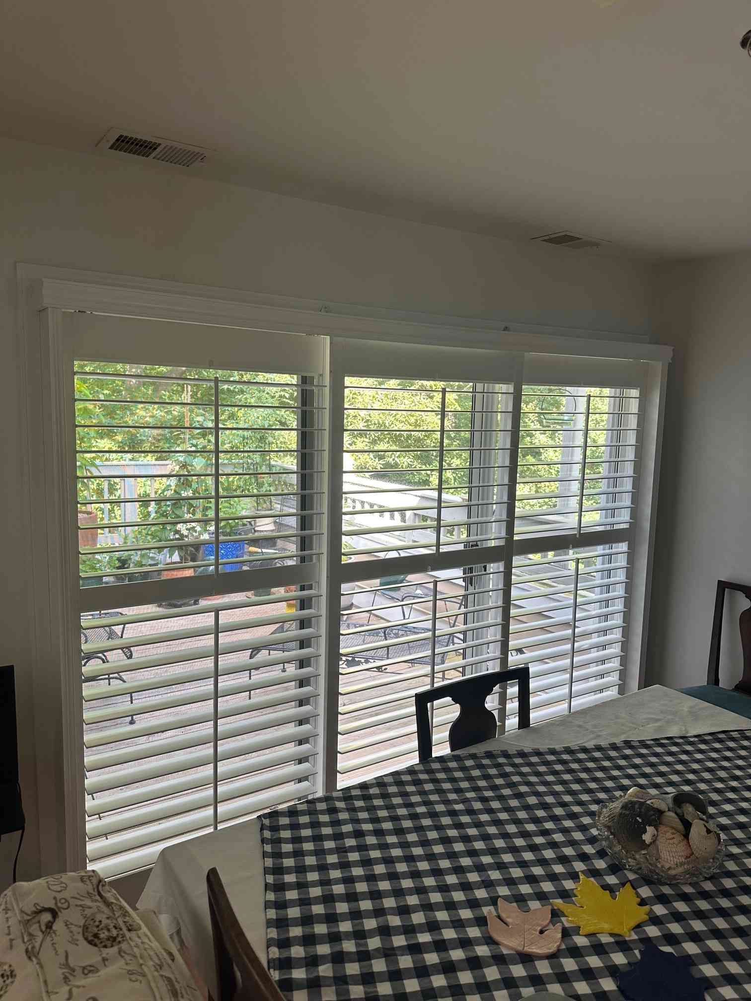 Dining room with a checkered tablecloth by large white plantation shutters overlooking greenery.