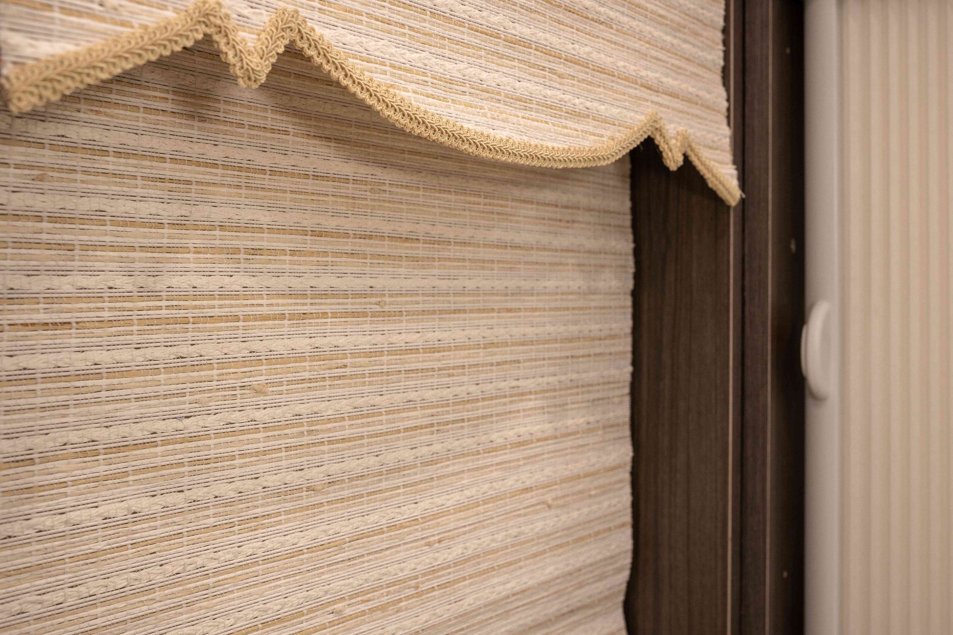 Close-up of beige woven fabric blinds with a scalloped valance beside a dark window frame