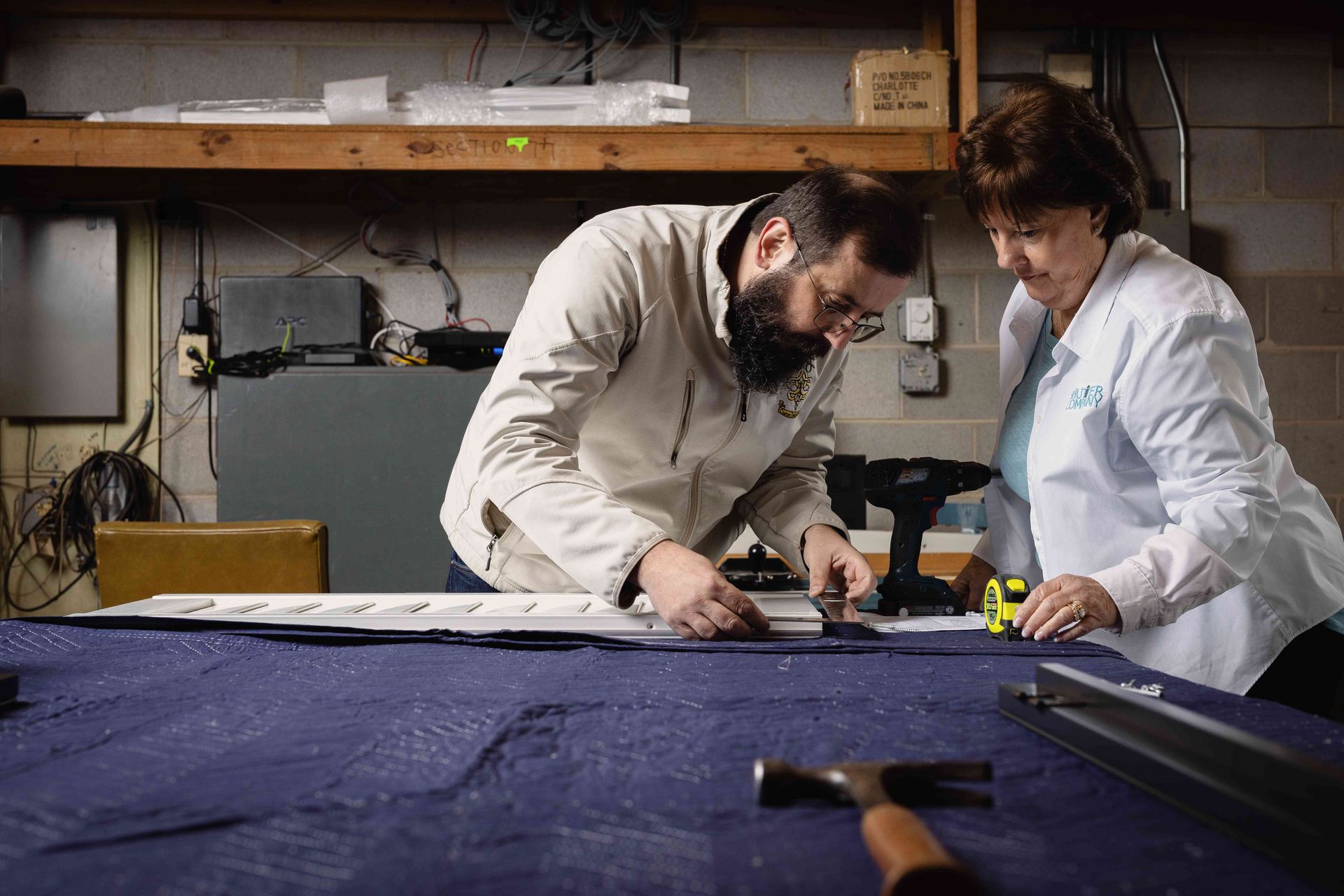 Two people in white coats measuring fabric on a large worktable in a workshop