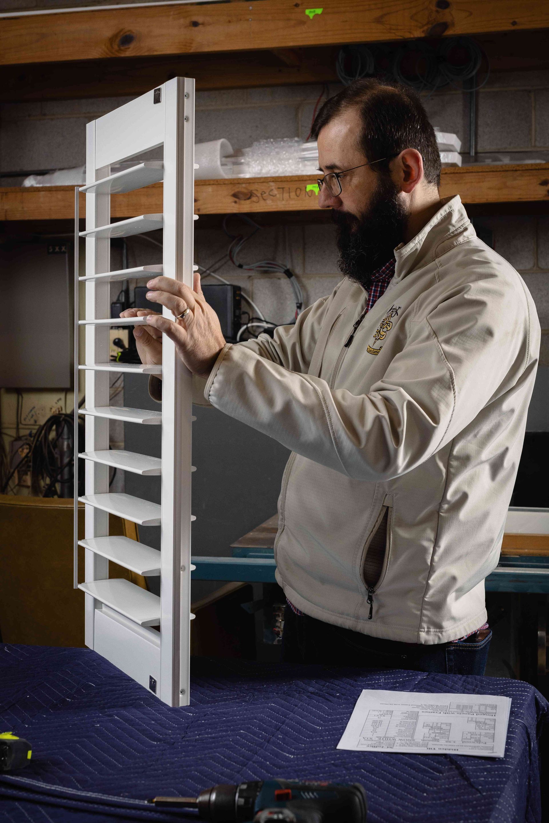 Man adjusting a white window shutter in a workshop, beside a table with papers
