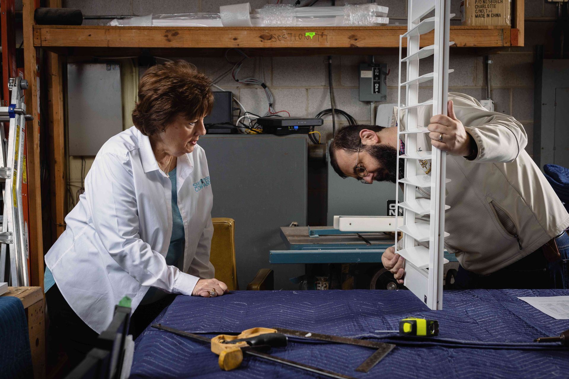 Two people measuring a white slatted panel on a workbench in a workshop.