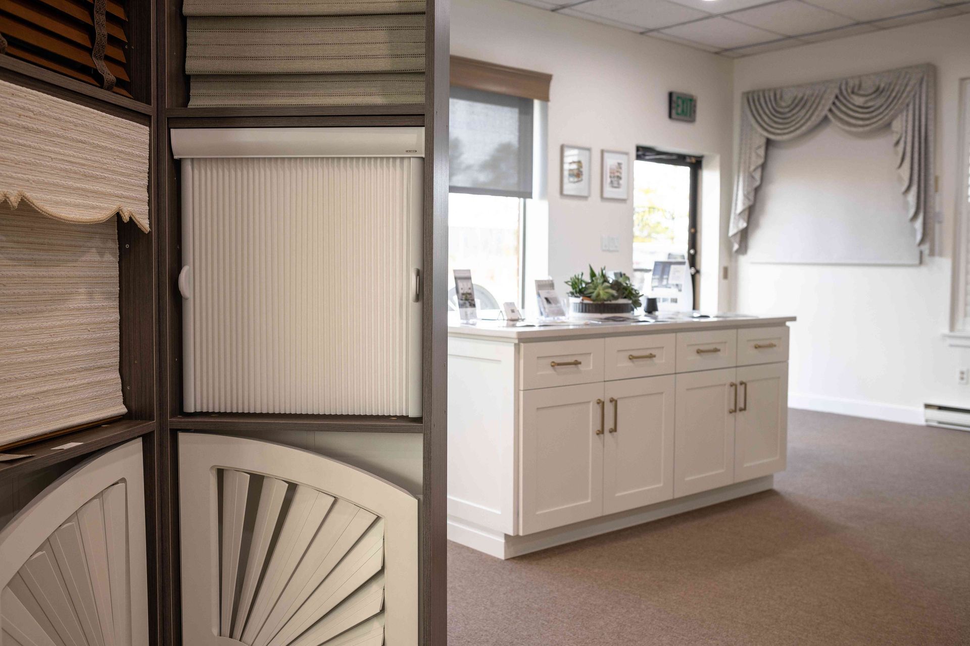 Display room with window treatments, white cabinetry, beige carpet, and sample blinds on the left.