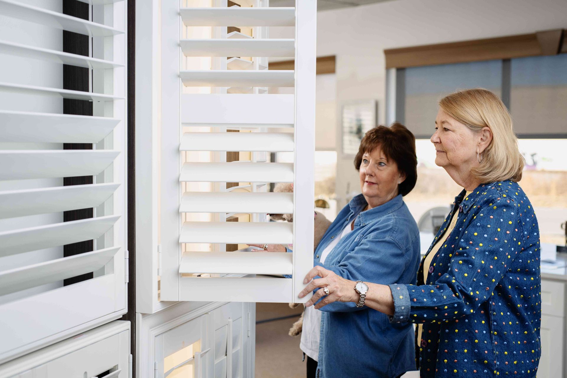 Two women examining white window shutters in a showroom