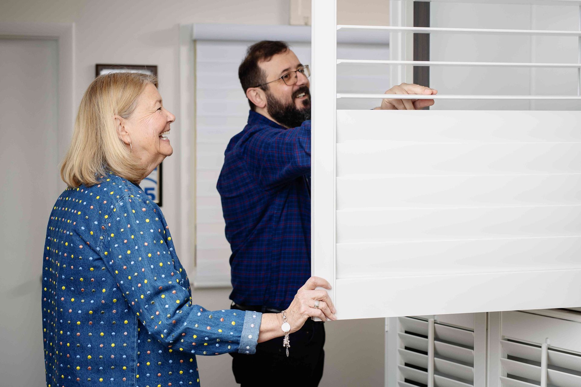 Two people opening white plantation shutters in a bright room.