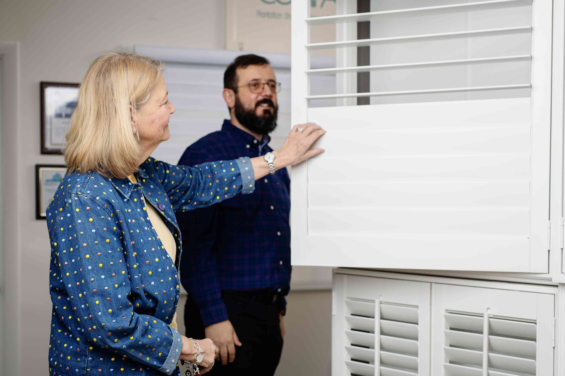Two people examining a white window blind indoors, with one hand touching the slats.