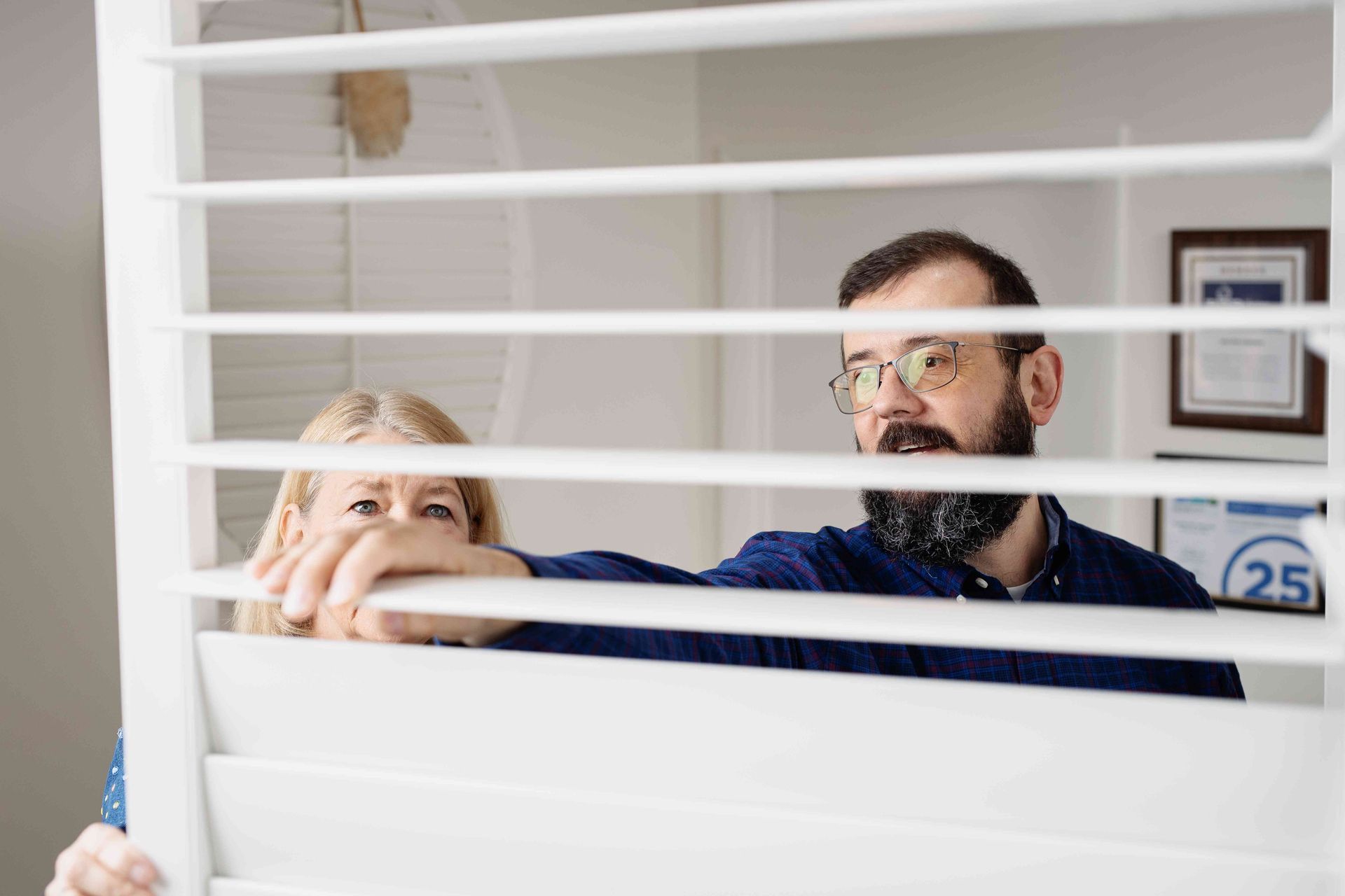 Two people look through white blinds in an office, one holding a blind open.