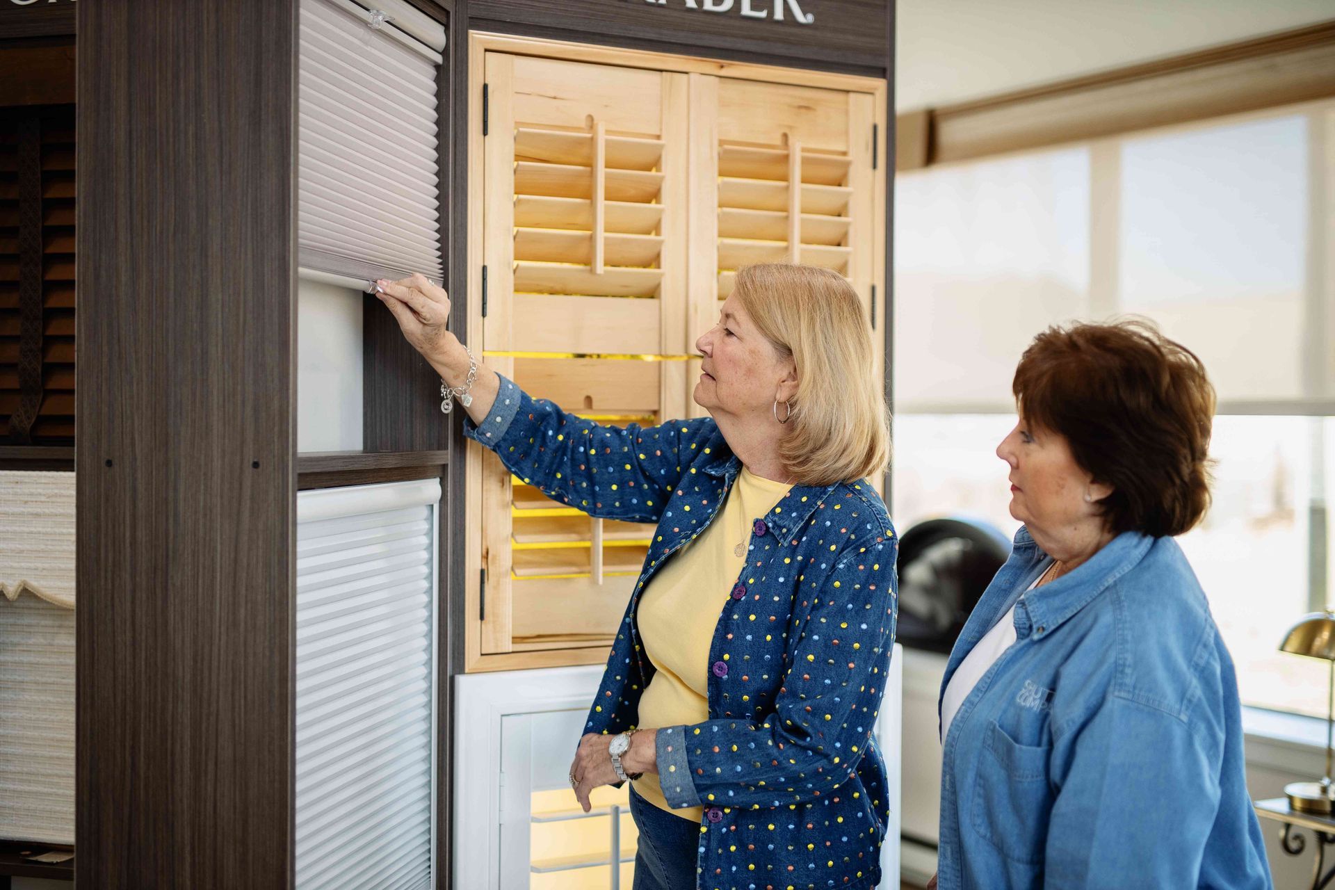 Two women comparing window blinds in a showroom, one touching a fabric sample.