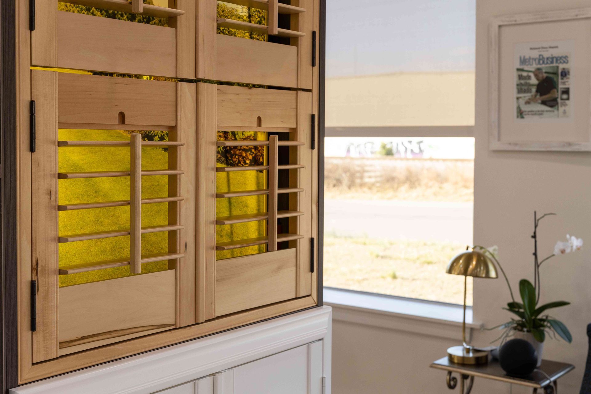 Sunlit room with wooden shutters, a window, and a small table holding a lamp and orchid.