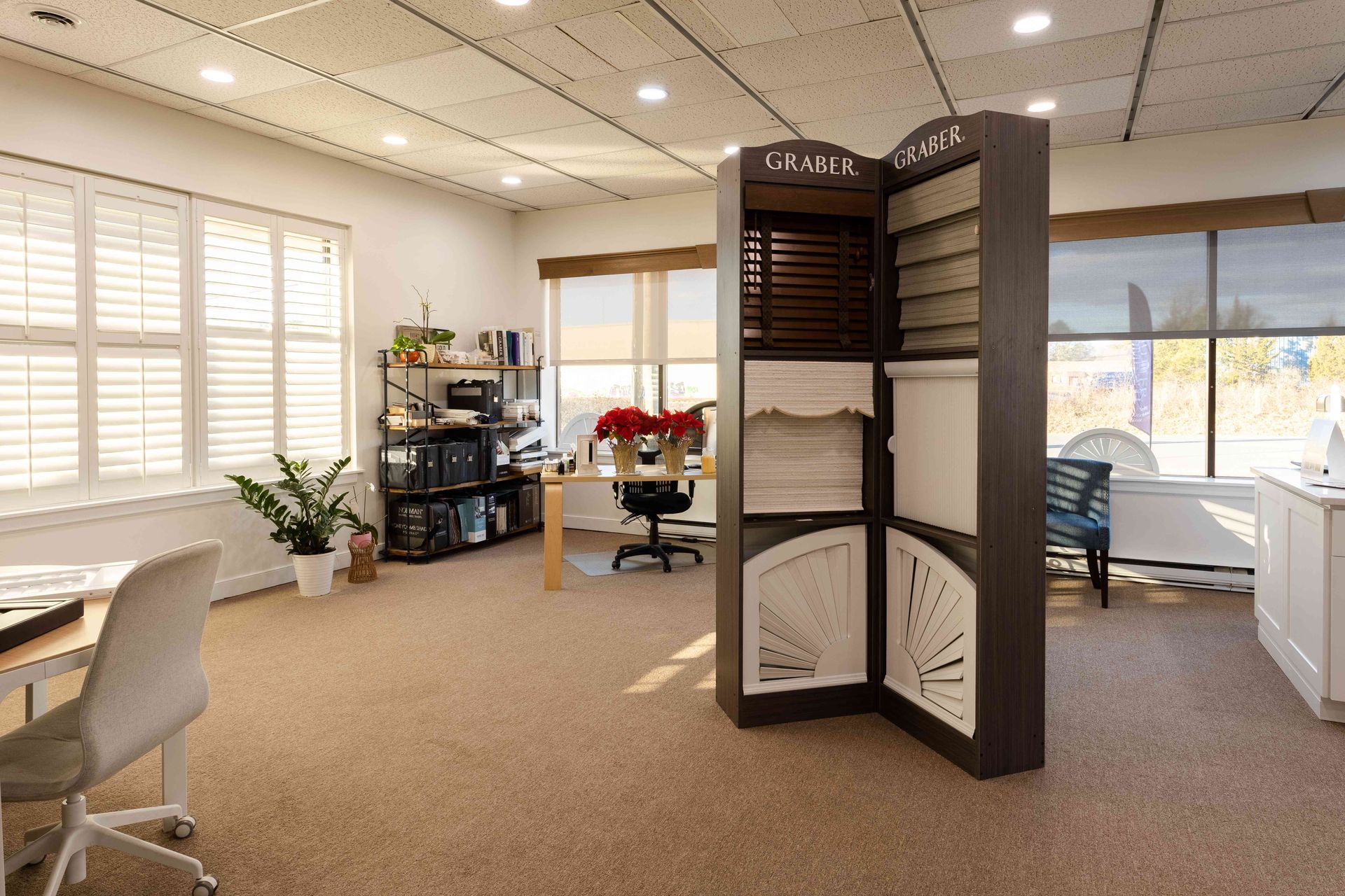 Bright office lounge with beige carpet, plants, chairs, desks, and a dark folding privacy screen near windows
