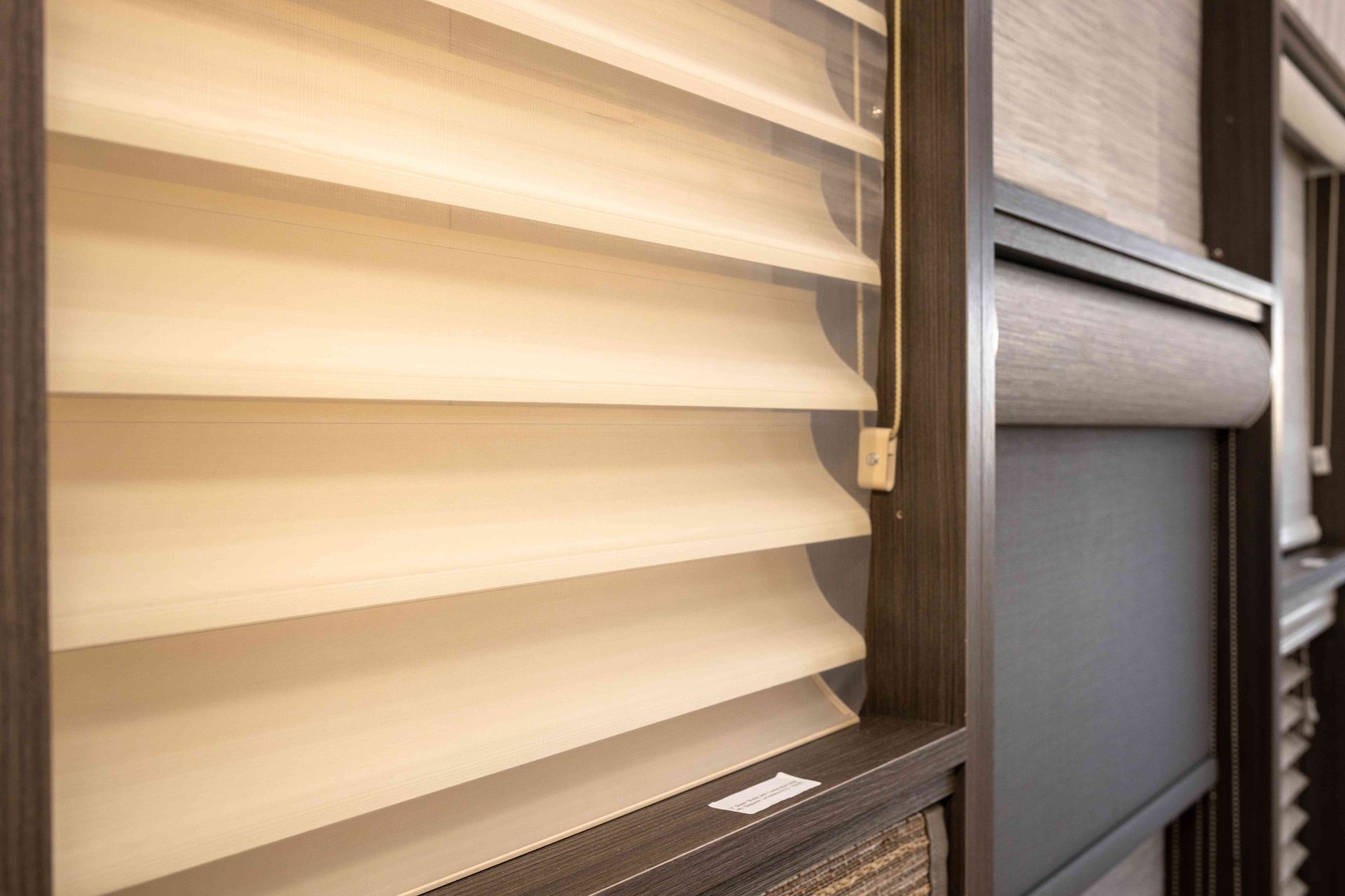Beige pleated window shade beside a dark wood window frame and gray blind