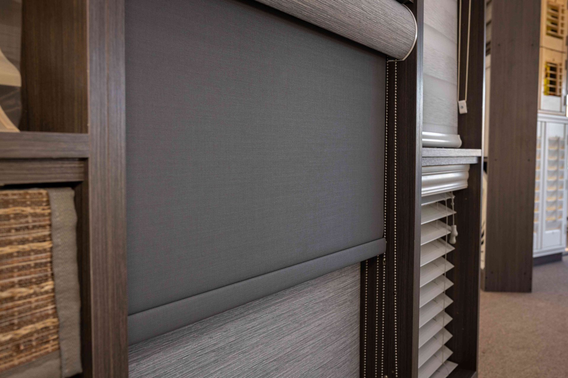 Gray roller window shade in a room display, with woven blinds and wood trim nearby