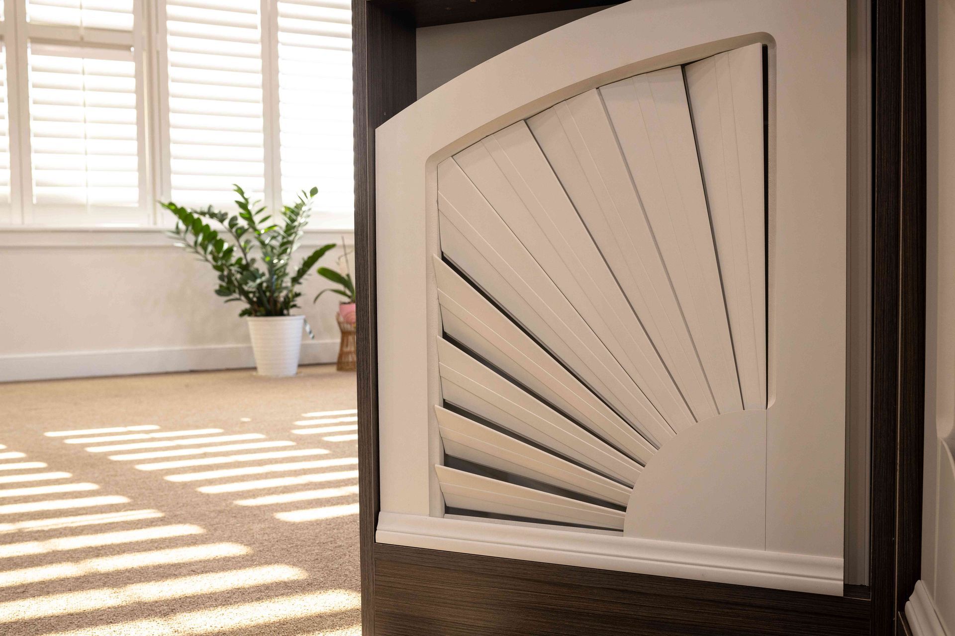 White decorative vent panel beside sunlit window with potted plant on a hardwood floor