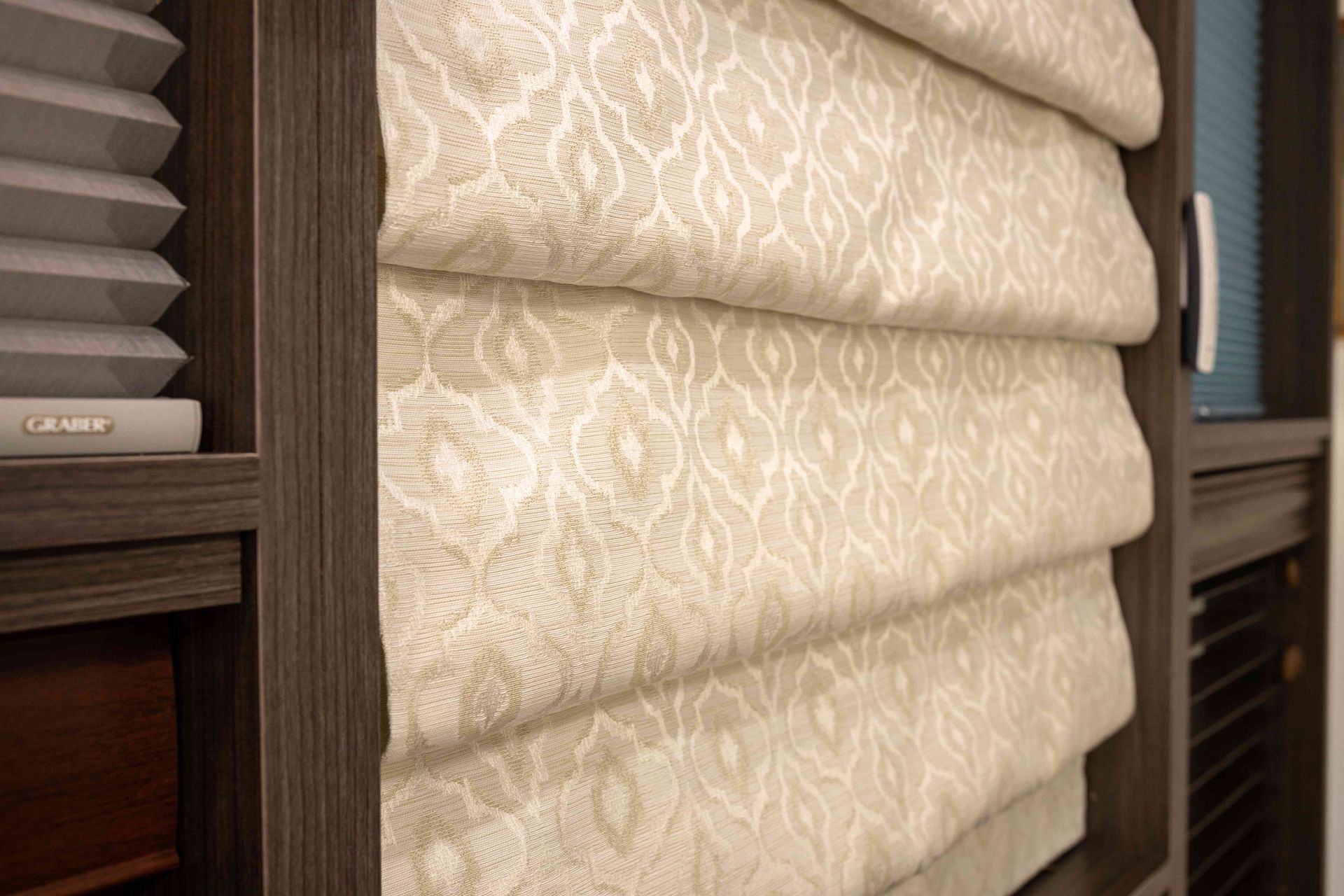 Beige patterned roman blinds in a wooden window frame with gray blinds beside them
