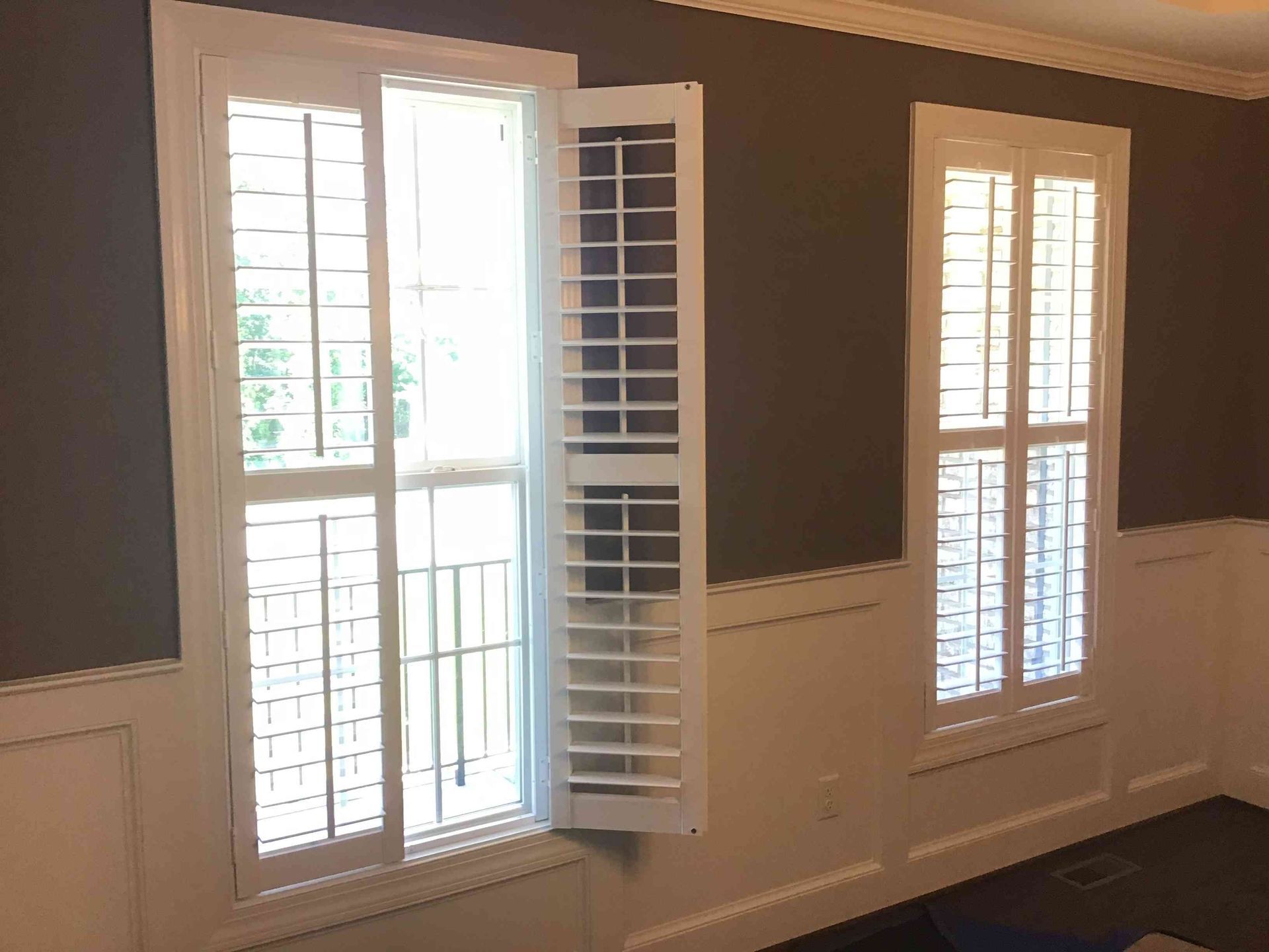 Empty room with brown walls, white wainscoting, and two sunlit windows with shutters.