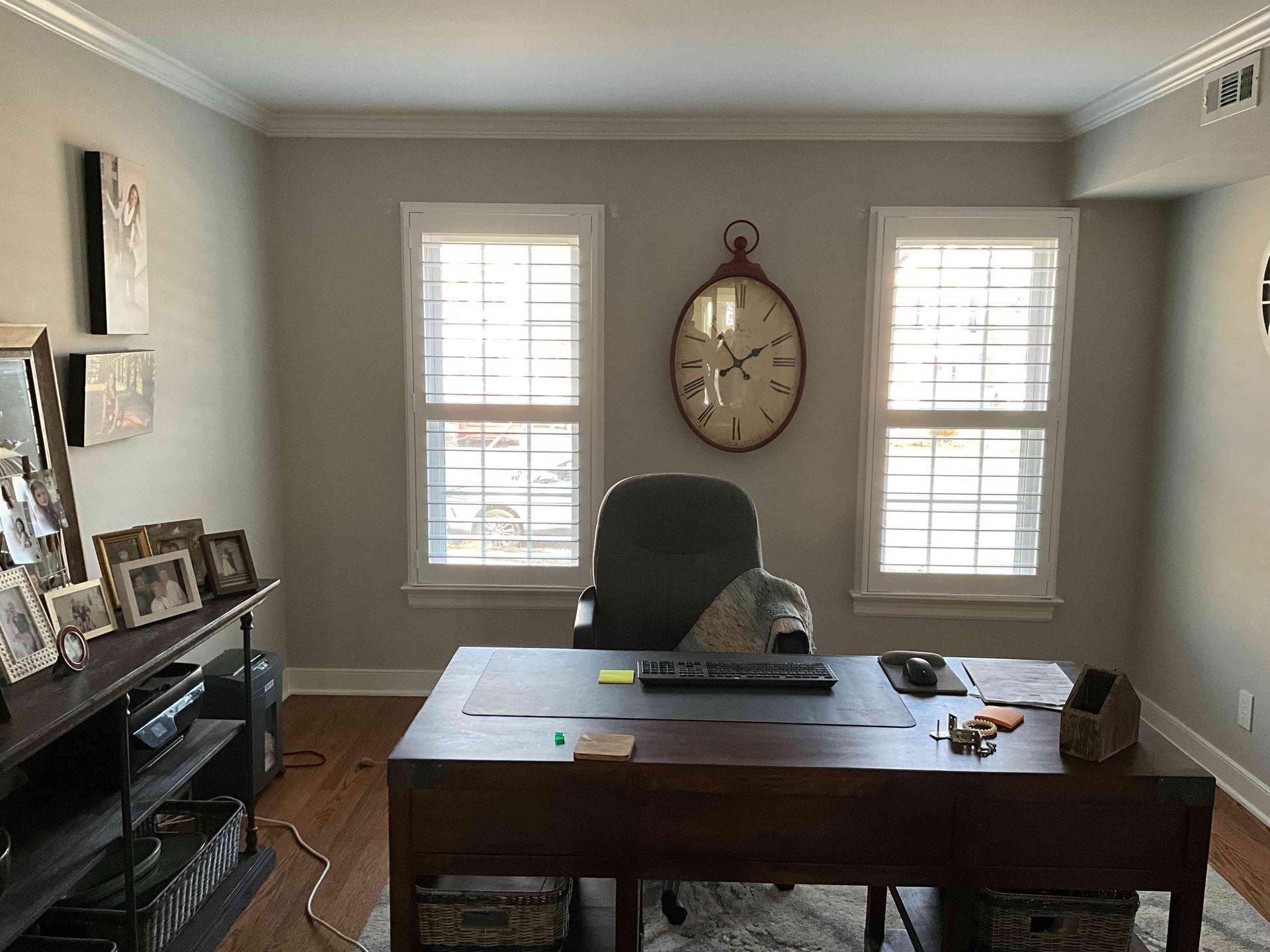 Home office with a wooden desk, chair, framed photos, and wall clock between two windows.