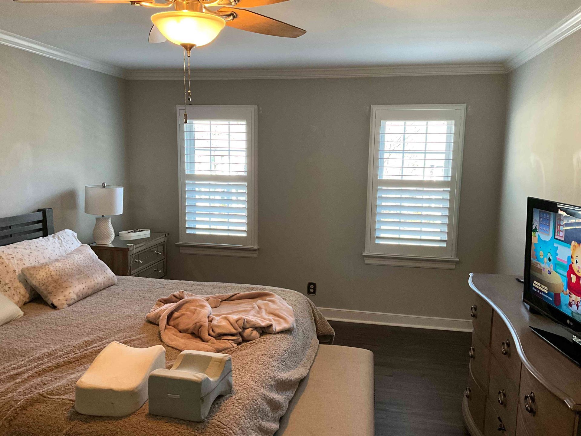 Neatly made bedroom with bed, dresser, TV, and two windows under a ceiling fan.