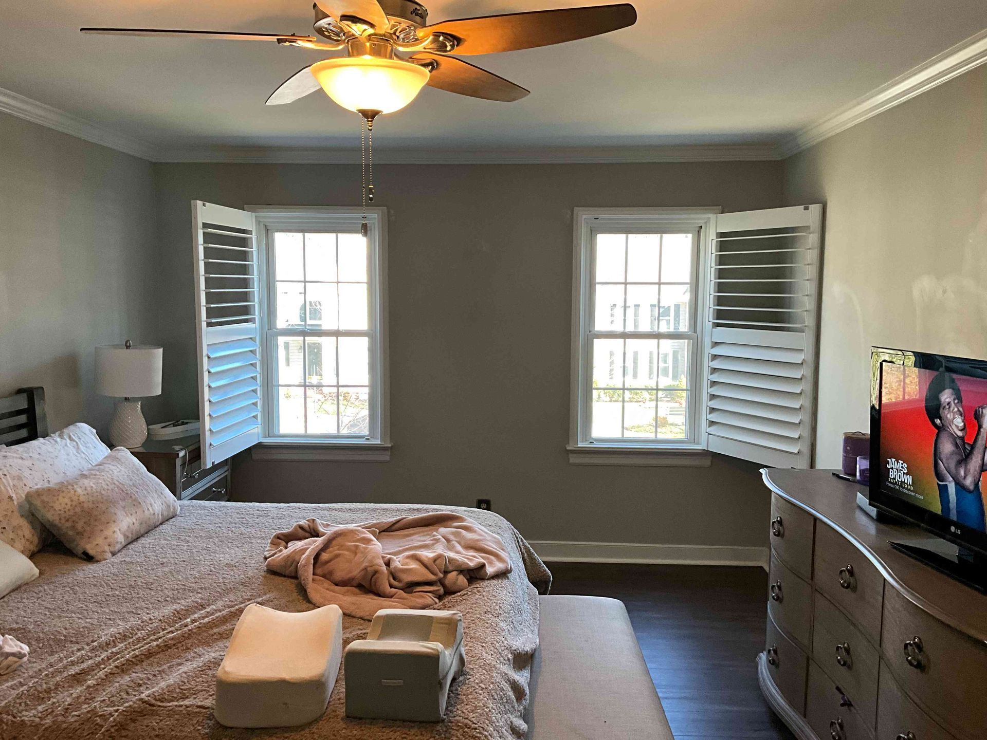 Bedroom with a bed, dresser, TV, ceiling fan, and two windows with shutters.