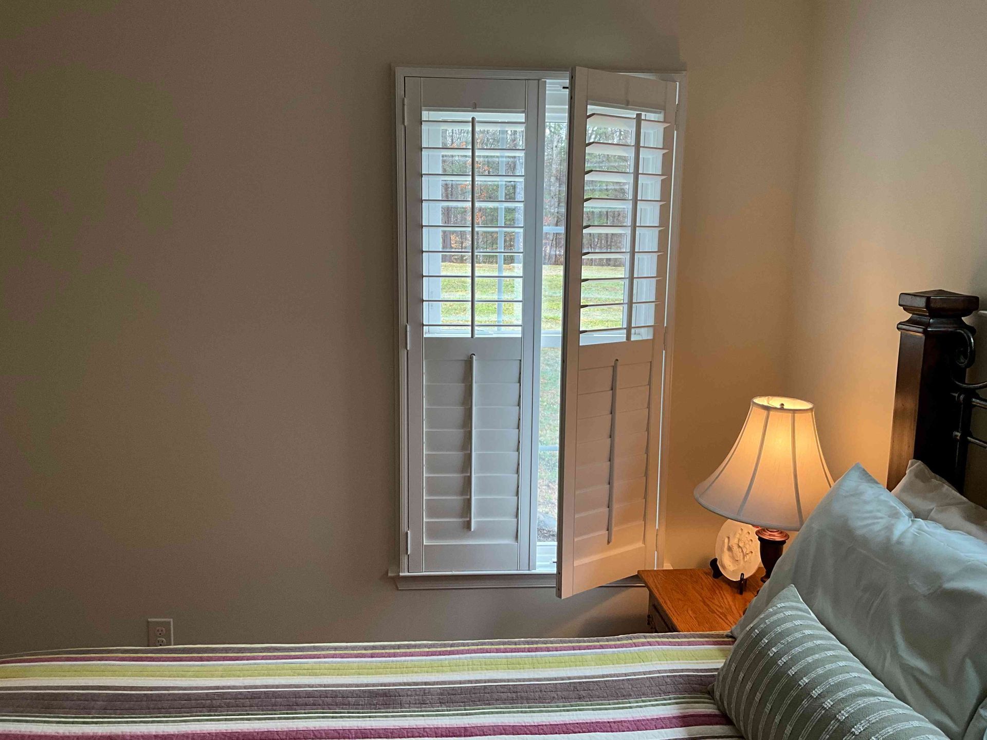 Bedroom with white shutters, bedside lamp, and a bed with striped bedding near a window.