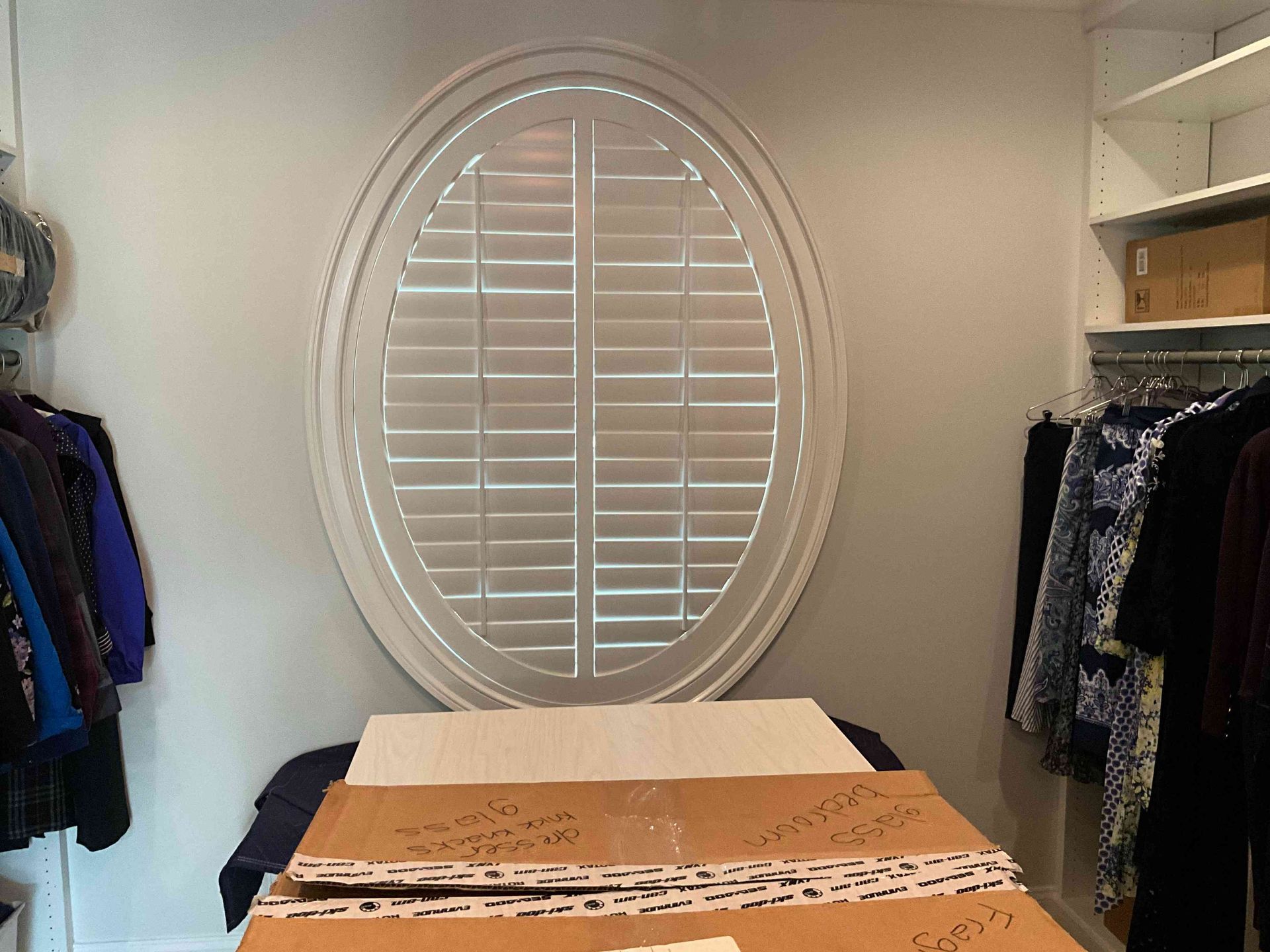 Walk-in closet with oval window, shelves of folded clothes, and clothing racks on both sides.