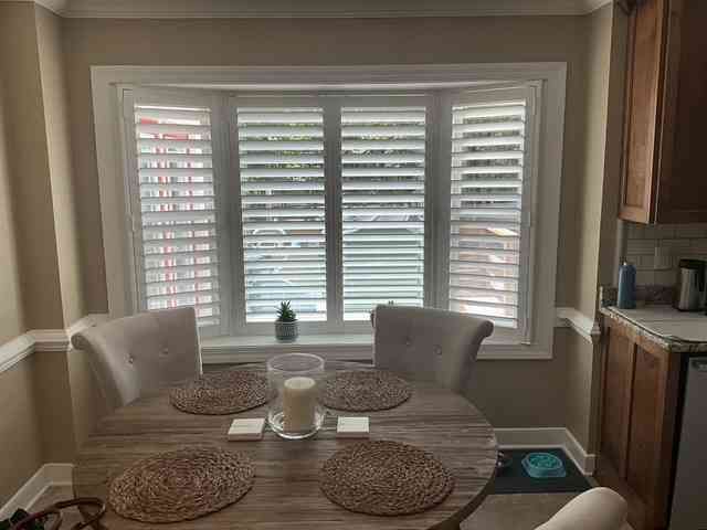Dining nook with round table, four chairs, and a window with white shutters and blinds.