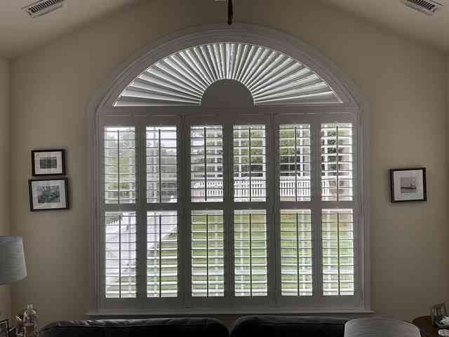 Arched window with white shutters and fanlight in a beige room, flanked by framed pictures.