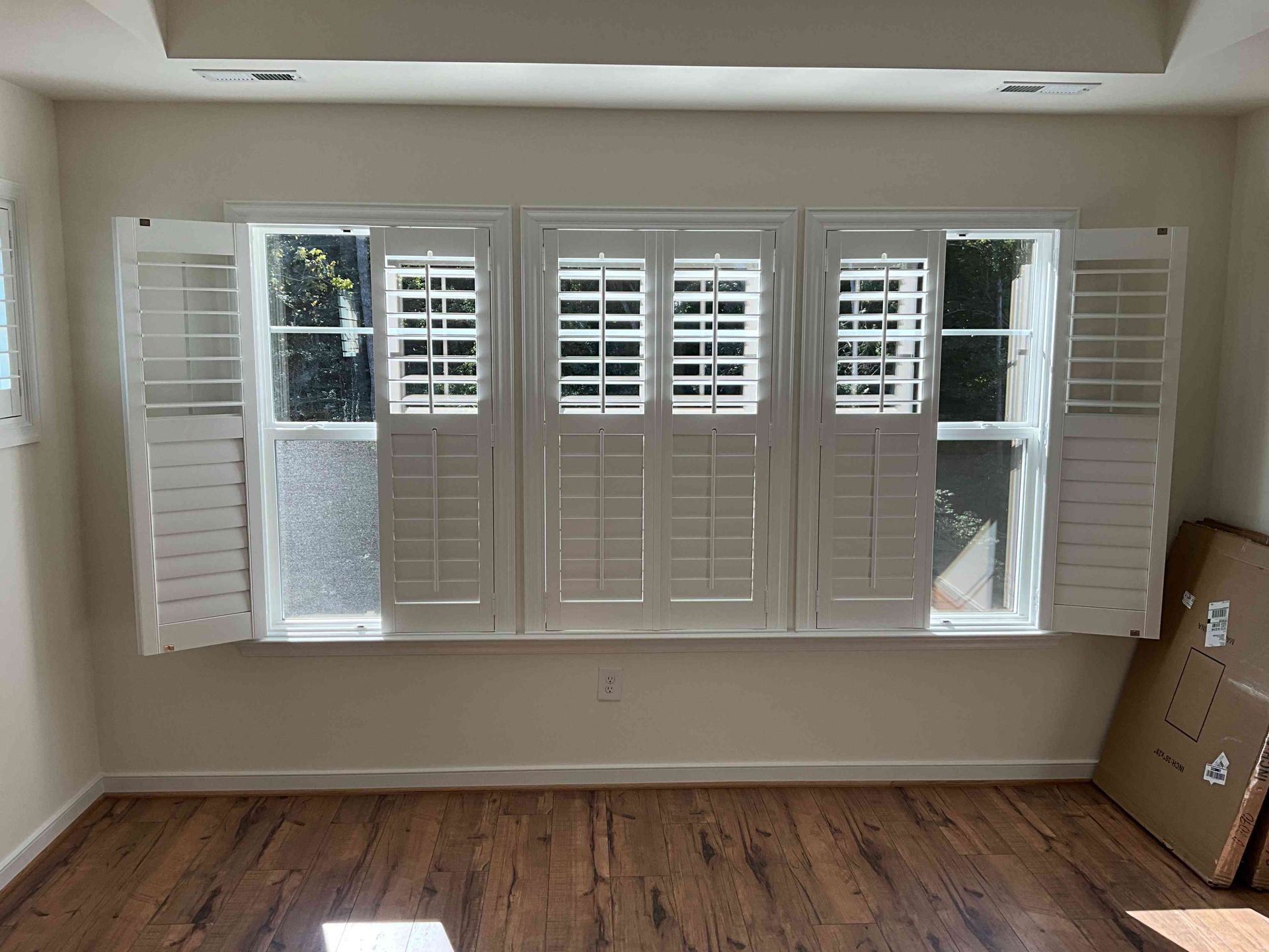 Empty room with a wide window, white shutters, and wood floor; cardboard boxes stacked at right.