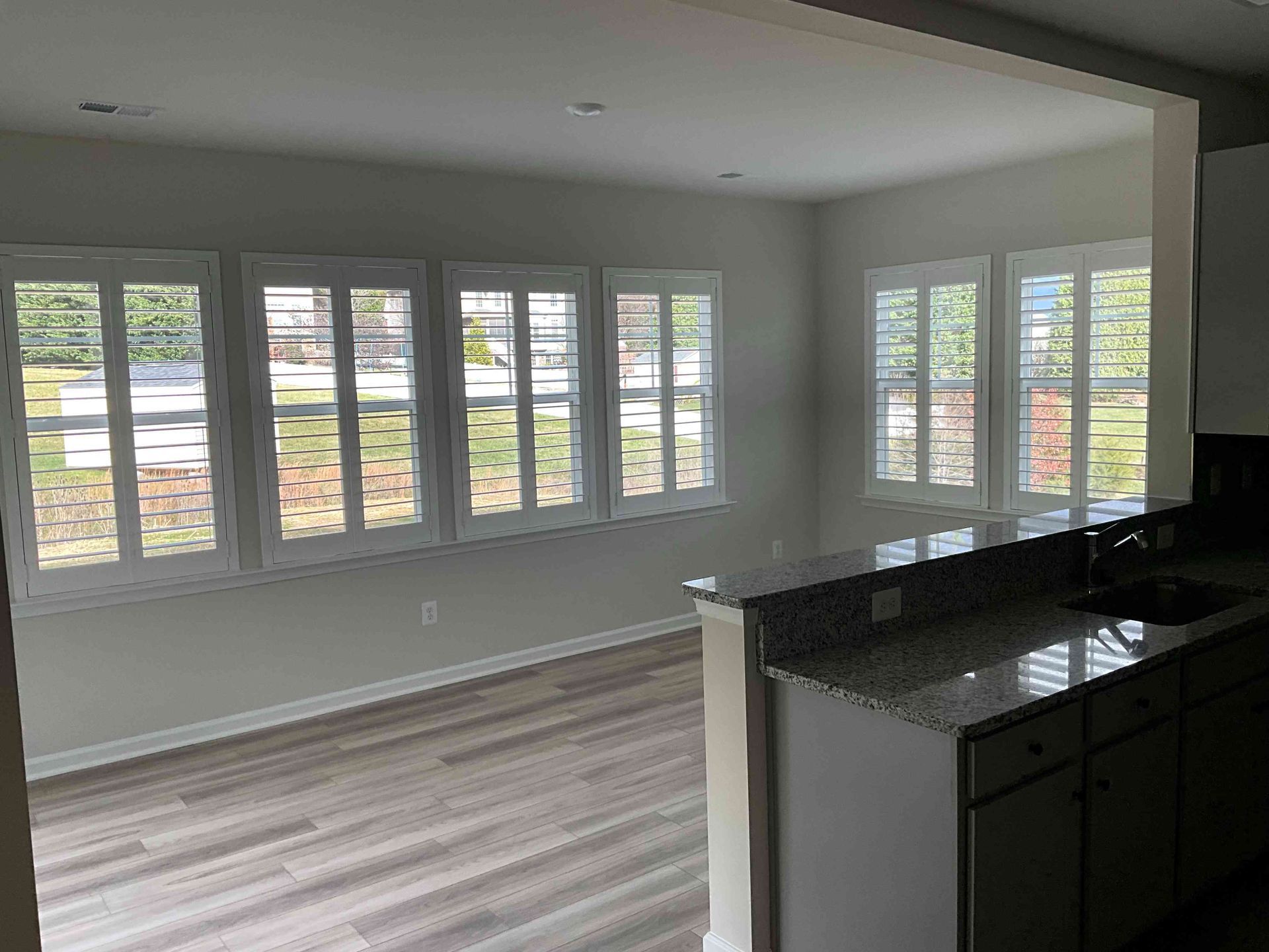 Empty modern living room with large windows, gray walls, light wood floors, and a dark granite kitchen island