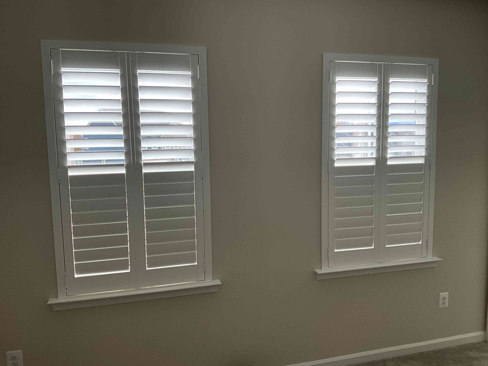 Empty beige room with two windows and white plantation shutters, one larger than the other.