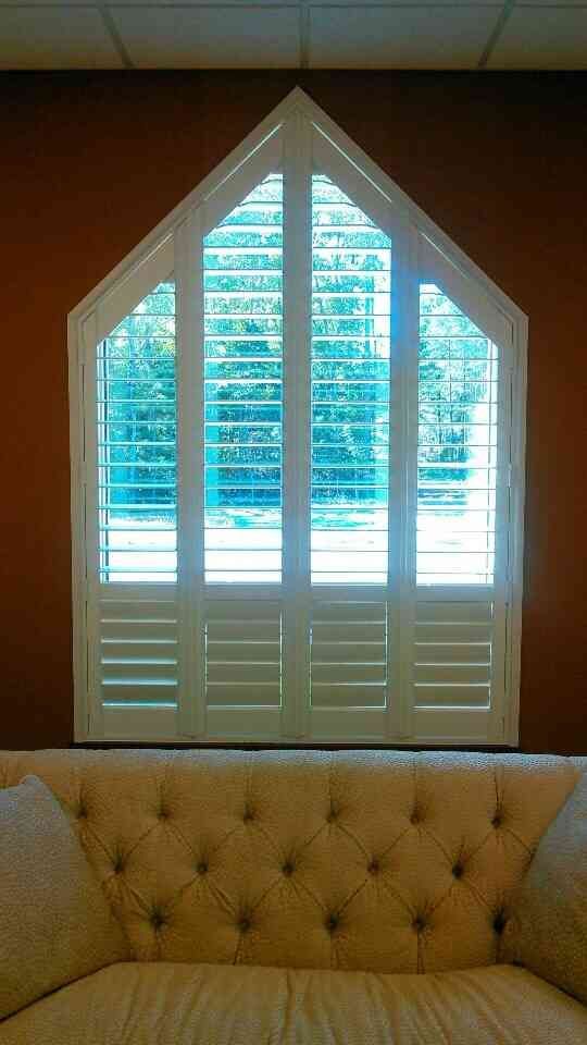 White arched window with shutters above a tufted beige bench against a brown wall, lit by daylight