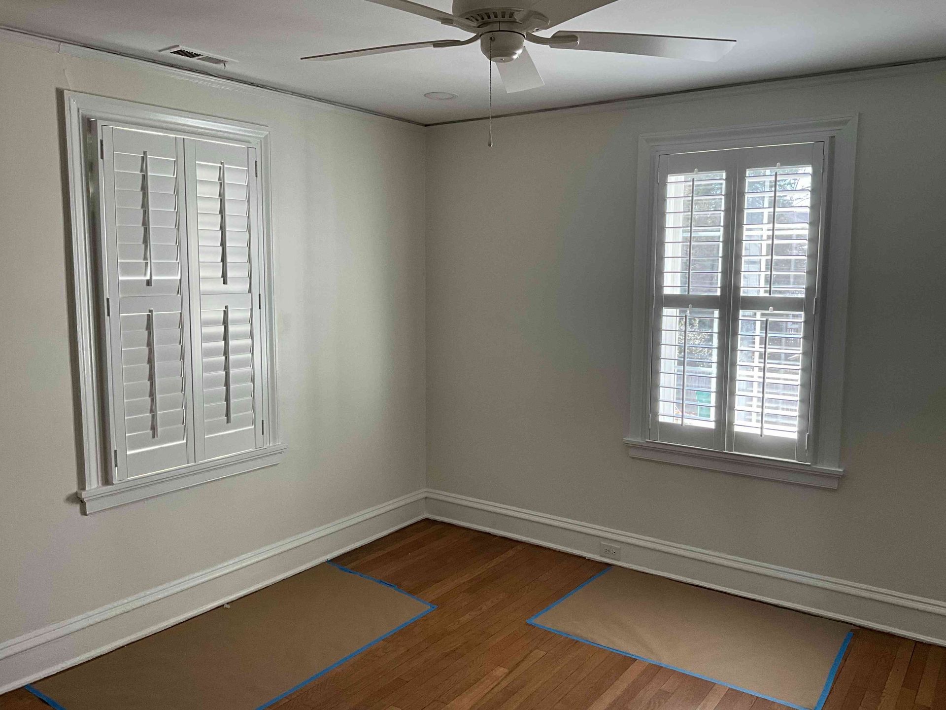 Empty beige bedroom corner with two shuttered windows, white trim, hardwood floor, and a ceiling fan