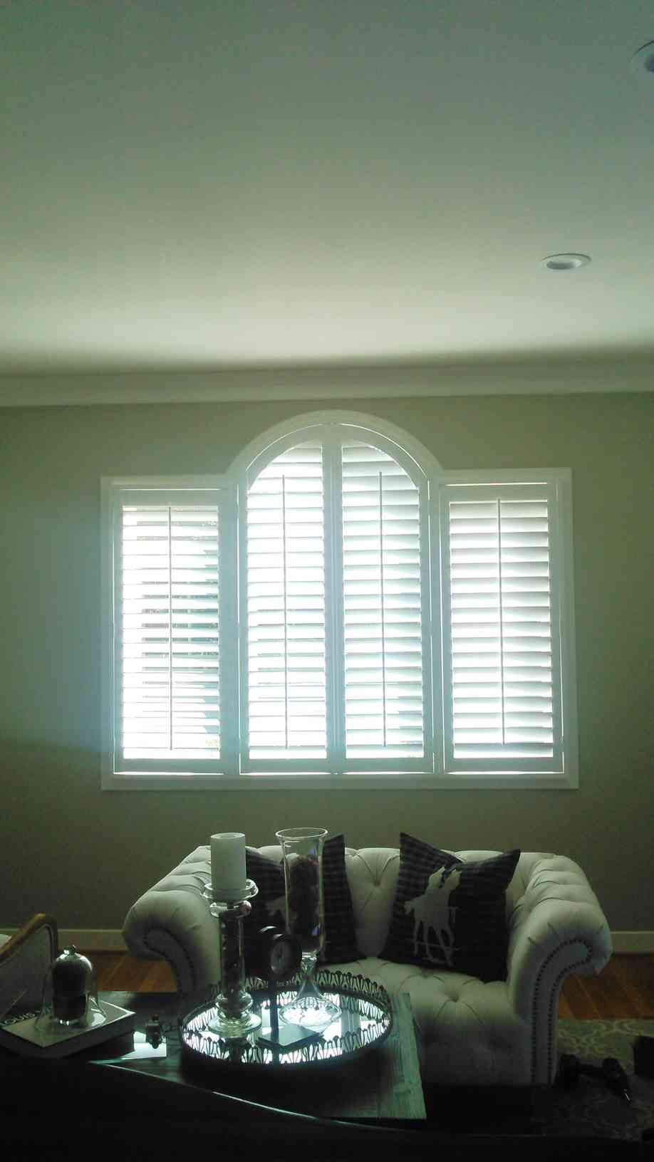 Living room with a white sofa and glass coffee table beneath a bright arched window with shutters