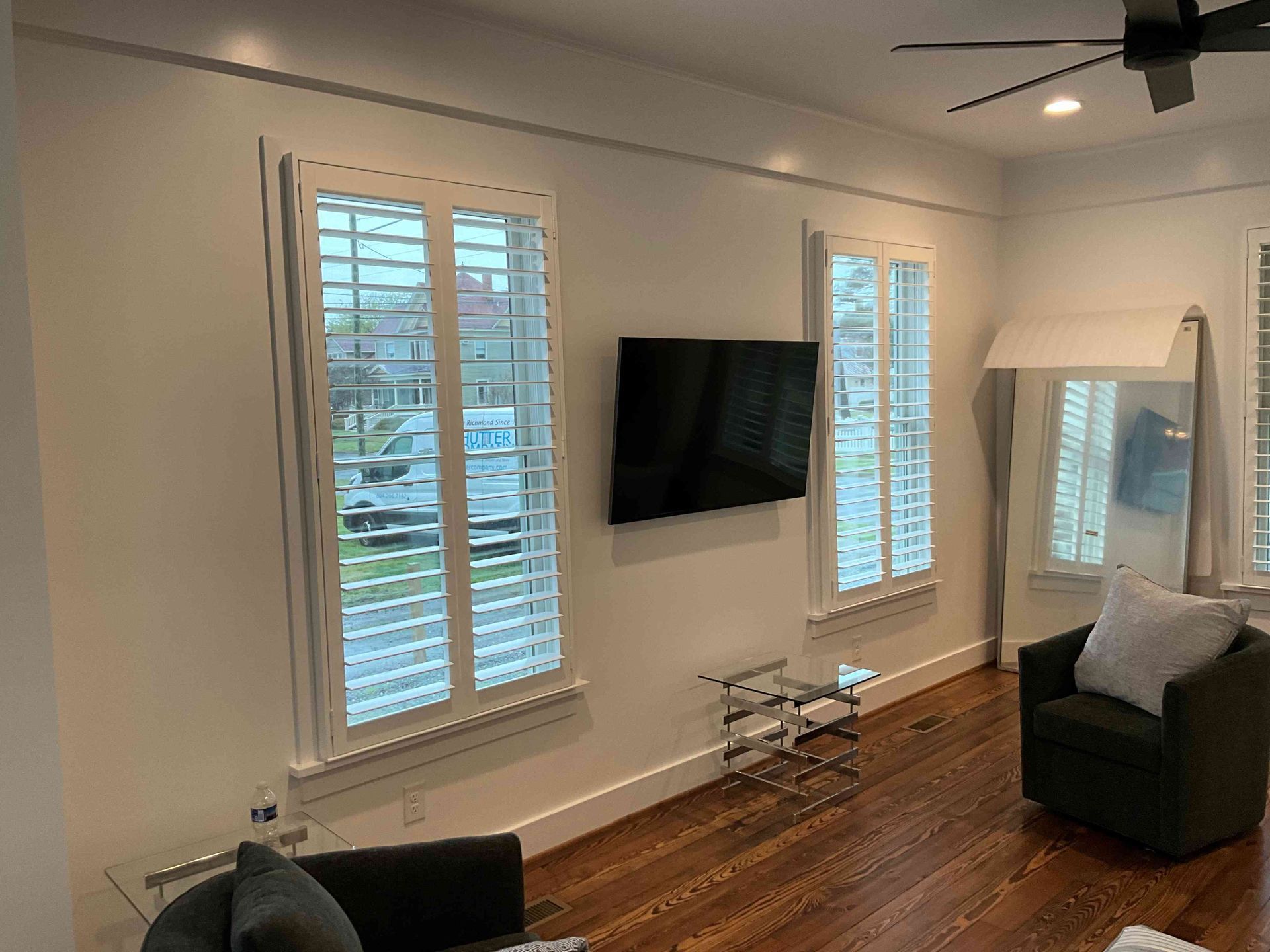 Modern living room with wall-mounted TV, white shutters, hardwood floor, and a gray armchair