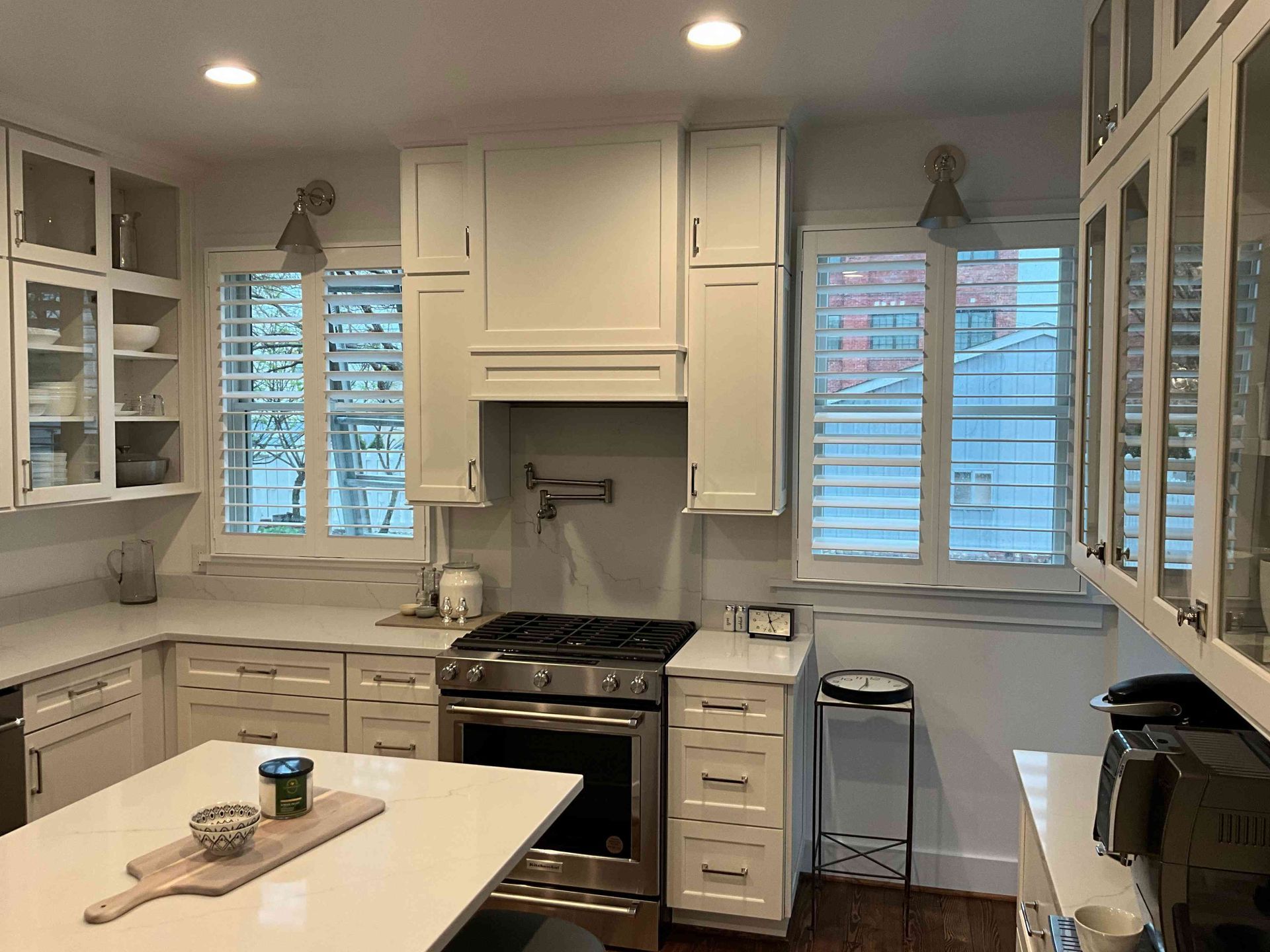 Bright white kitchen with central island, stainless stove, cabinets, and windows with shutters.