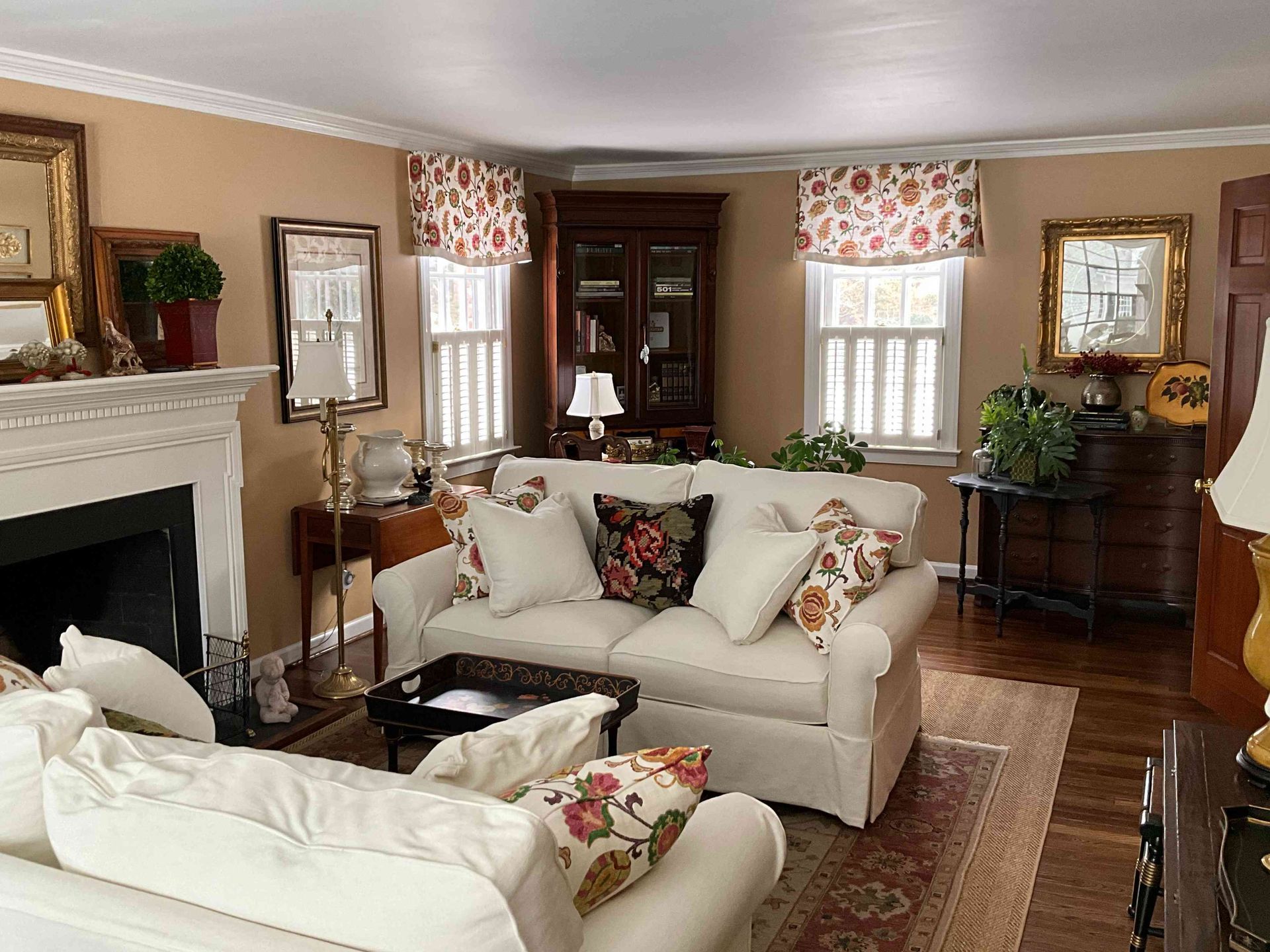 Cozy living room with cream sofas, fireplace, large windows, dark wood cabinets, and floral curtains.