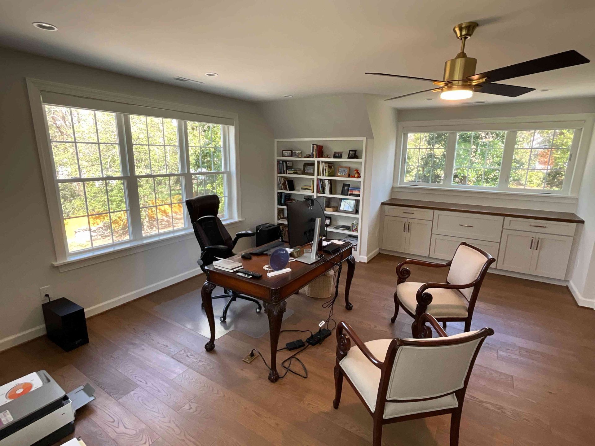 Bright home office with desk, chairs, bookshelf, windows, and ceiling fan in a sunlit room