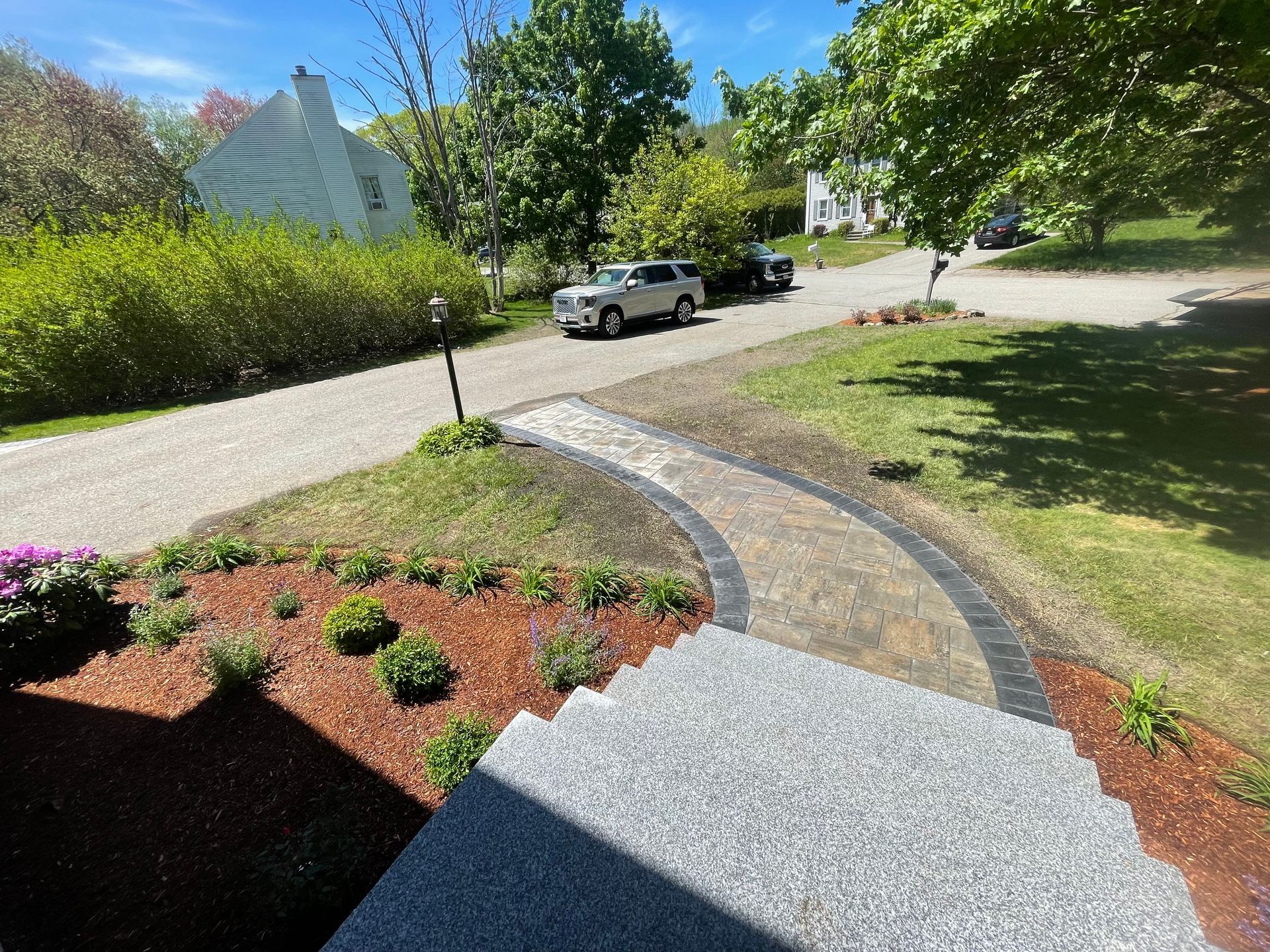 Curved stone path through a sunny garden with red mulch, shrubs, trees, and a parked white SUV nearby.