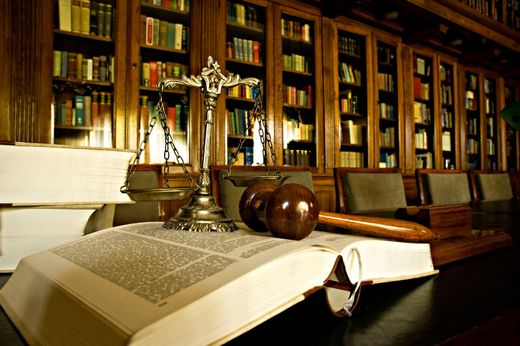 Open law book on desk in a library, with scales of justice and wooden gavel nearby.